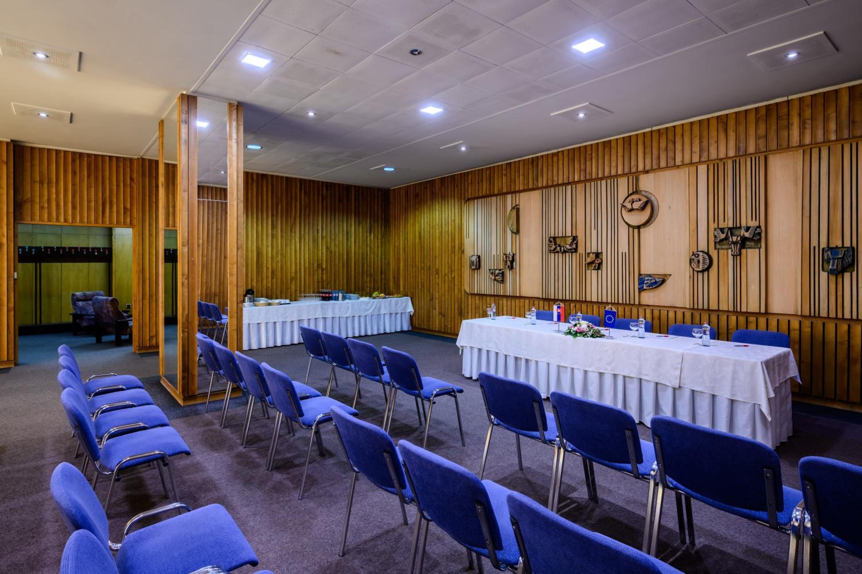 Ein Konferenzraum mit blauen Stühlen, einem langen Tisch mit weißen Tischdecken und Holzvertäfelungen an den Wänden.