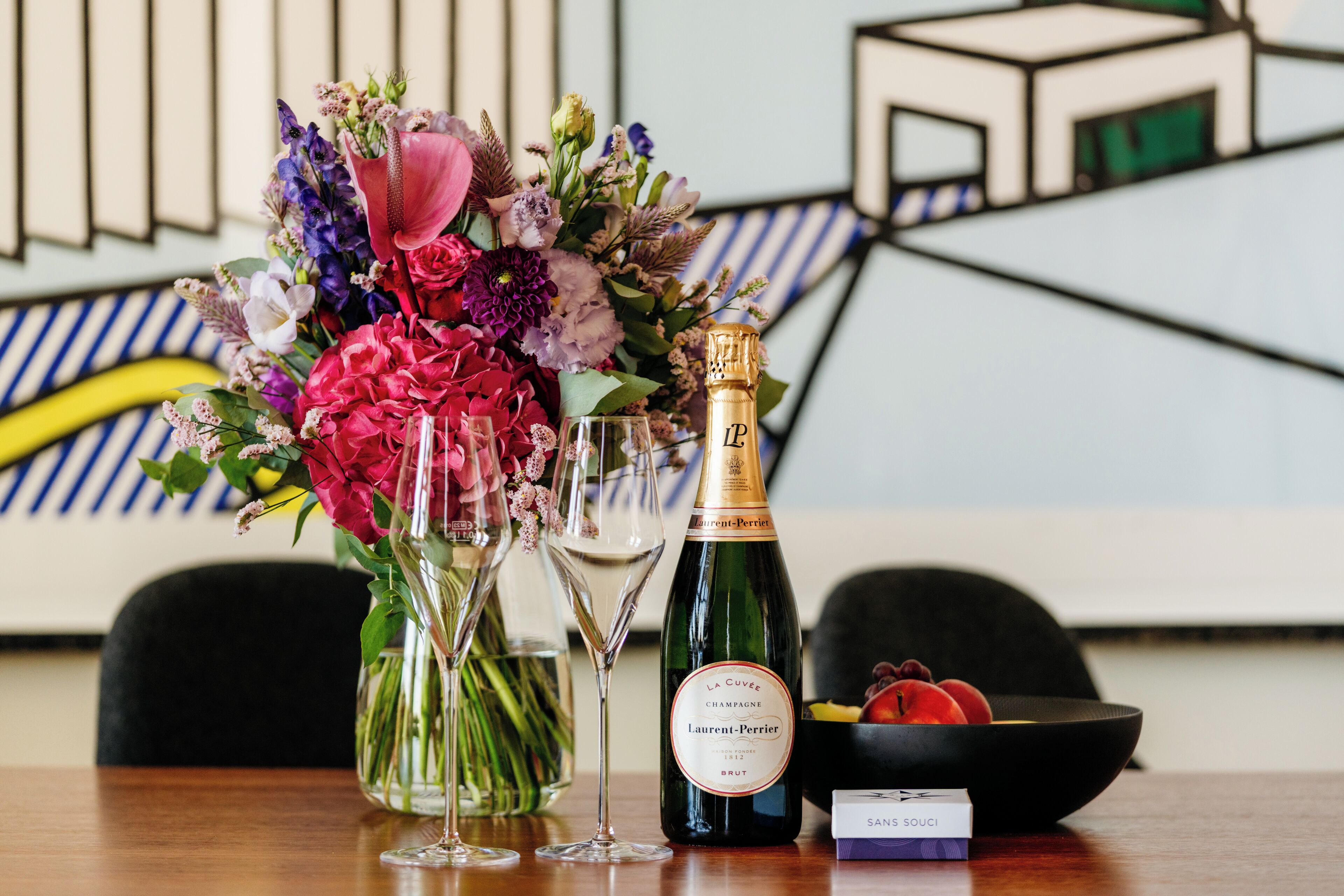A table with a bottle of champagne, two wine glasses, a floral arrangement, and a bowl of fruit.