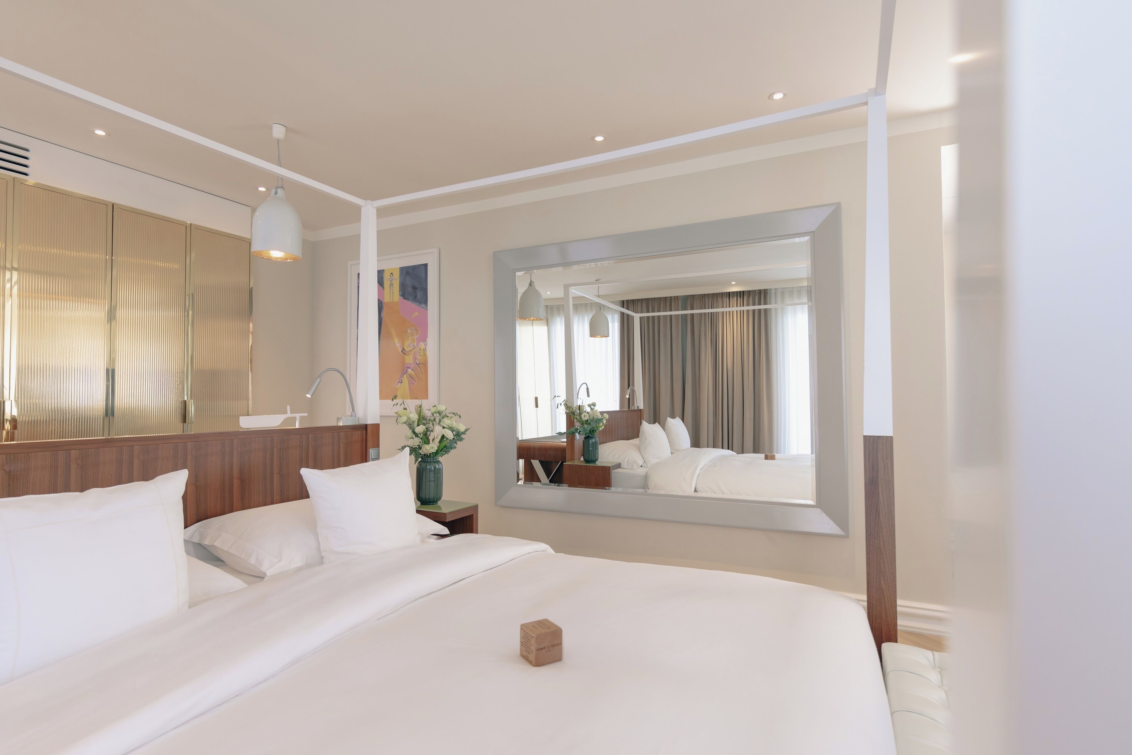 A modern hotel room with a large bed, a bedside table with a lamp, a mirror, and a view of the room through a window.