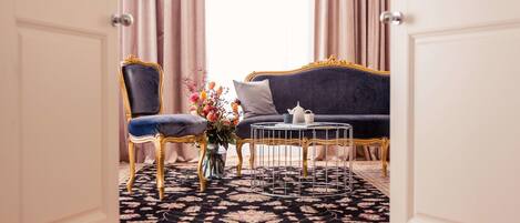 Une pièce avec un canapé bleu foncé, un fauteuil à cadre doré, une petite table ronde et un arrangement floral posé sur un tapis à motifs.