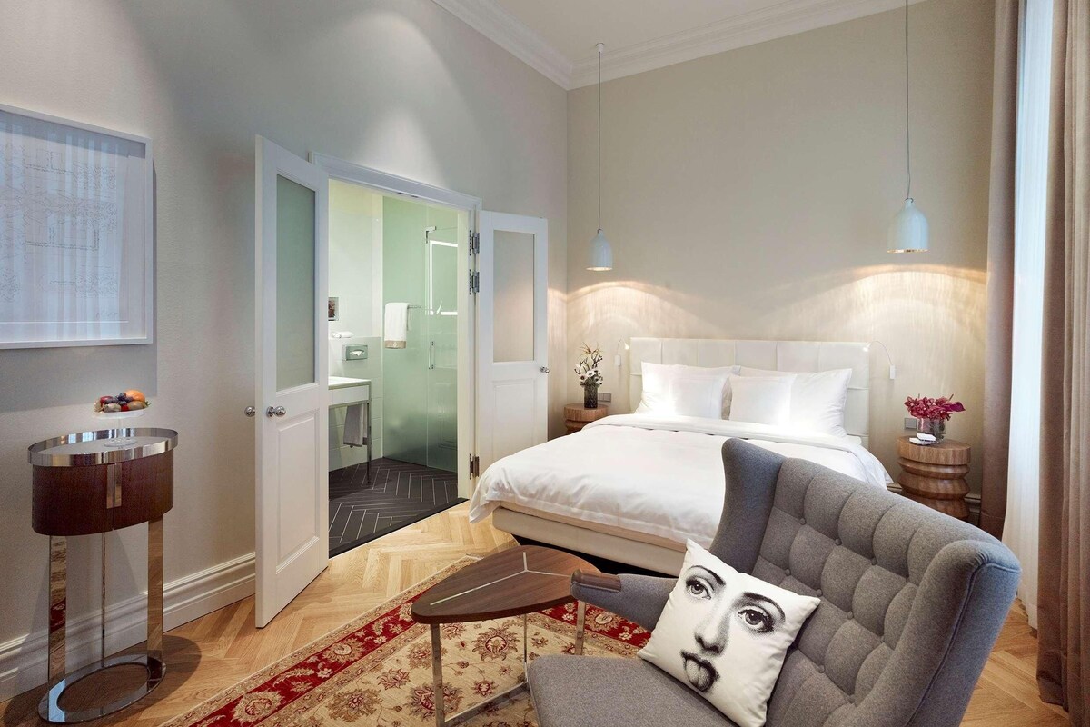 A hotel room with a bed, a chair, a bedside table, a lamp, a mirror, and a door leading to a bathroom.