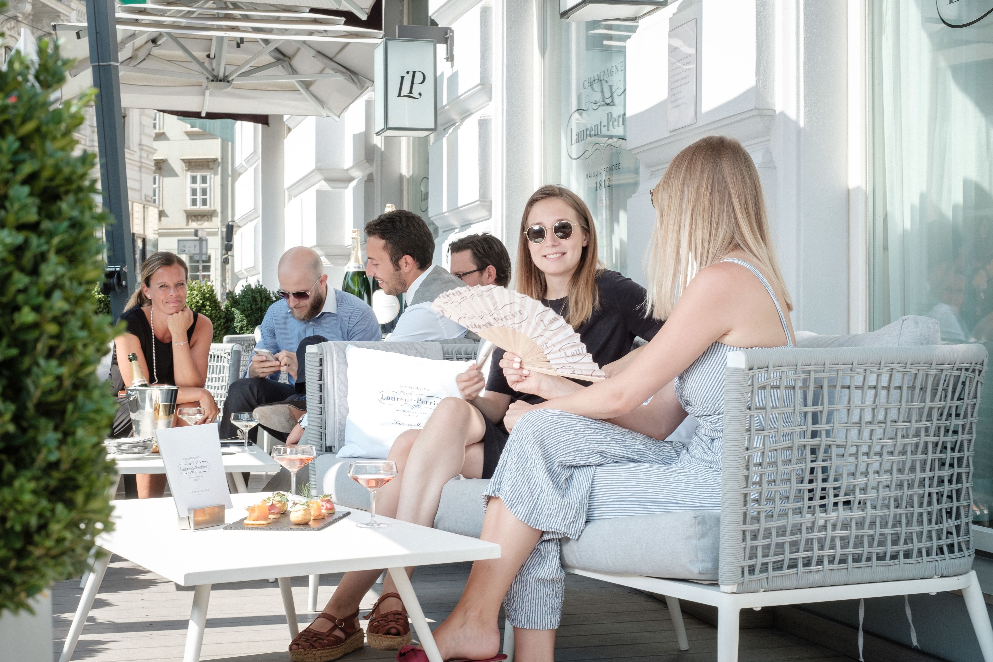 Un grupo de personas sentadas en un café al aire libre con mobiliario de mimbre, una carta y bebidas sobre la mesa.