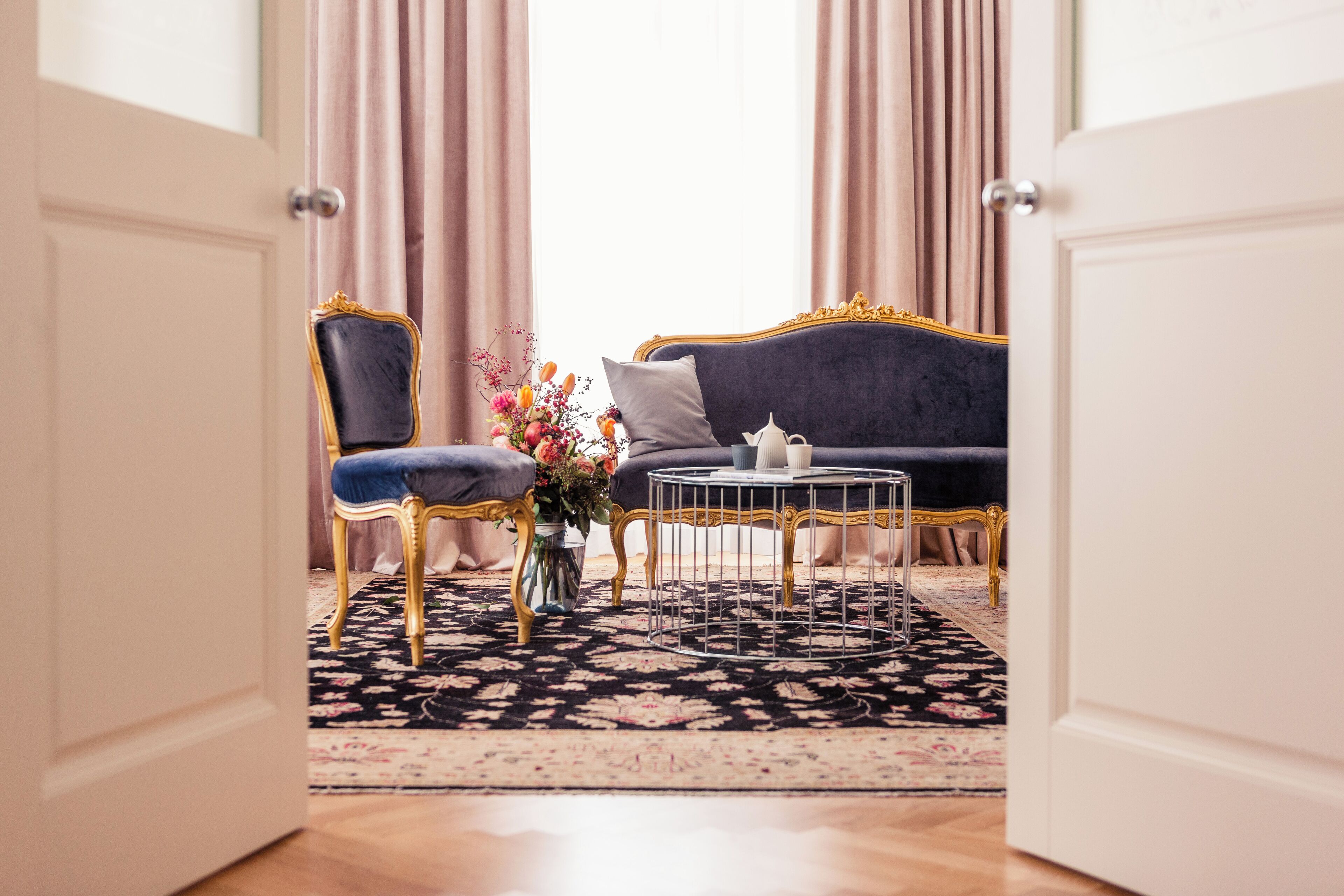 A room with a dark blue sofa, a gold-framed chair, a small round table, and a floral arrangement on a patterned rug.