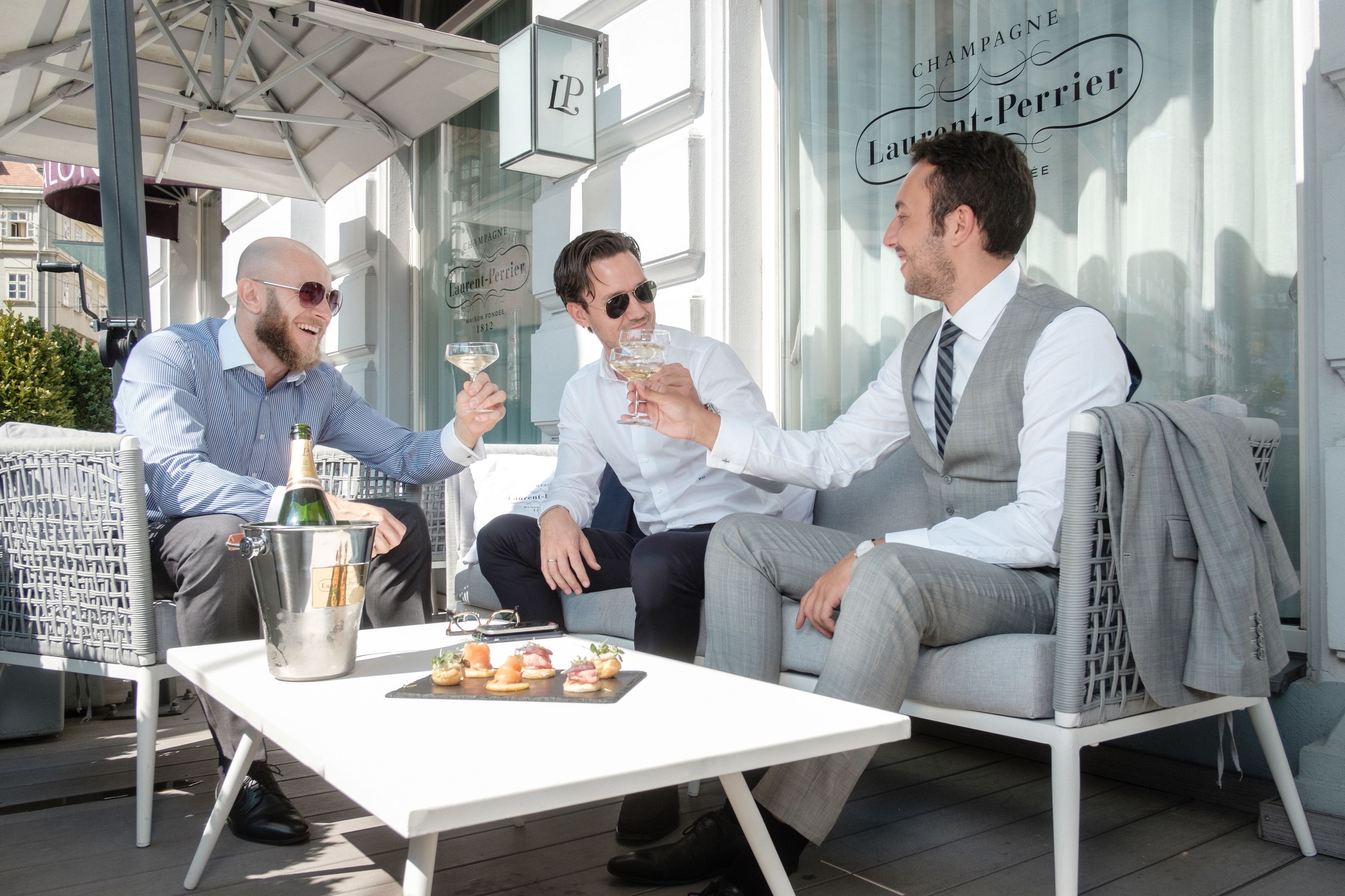 Three men seated at an outdoor table with drinks and appetizers, in a setting with a view of a building with signage.