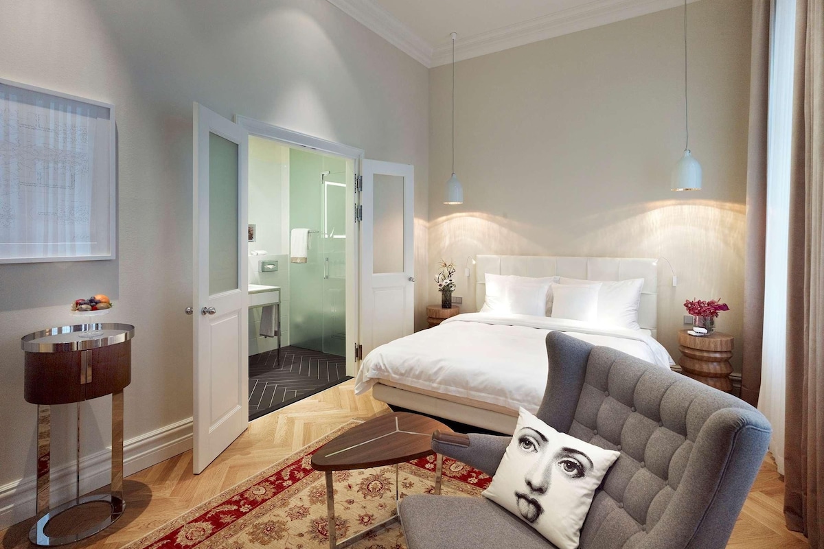 A hotel room with a bed, a chair, a bedside table, a lamp, a mirror, and a door leading to a bathroom.