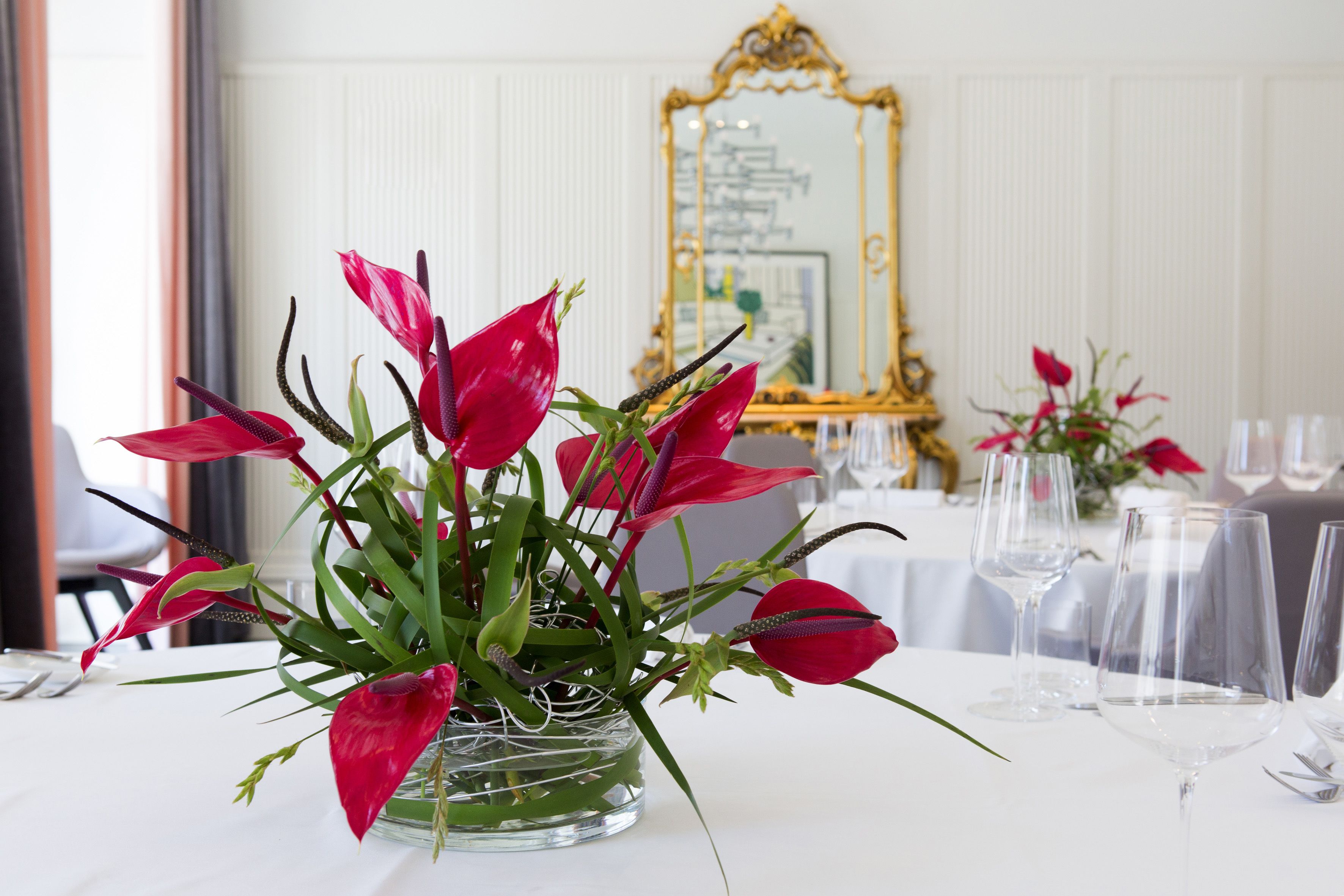 A dining table with a clear glass vase containing red flowers, a gold-framed mirror, and empty wine glasses.