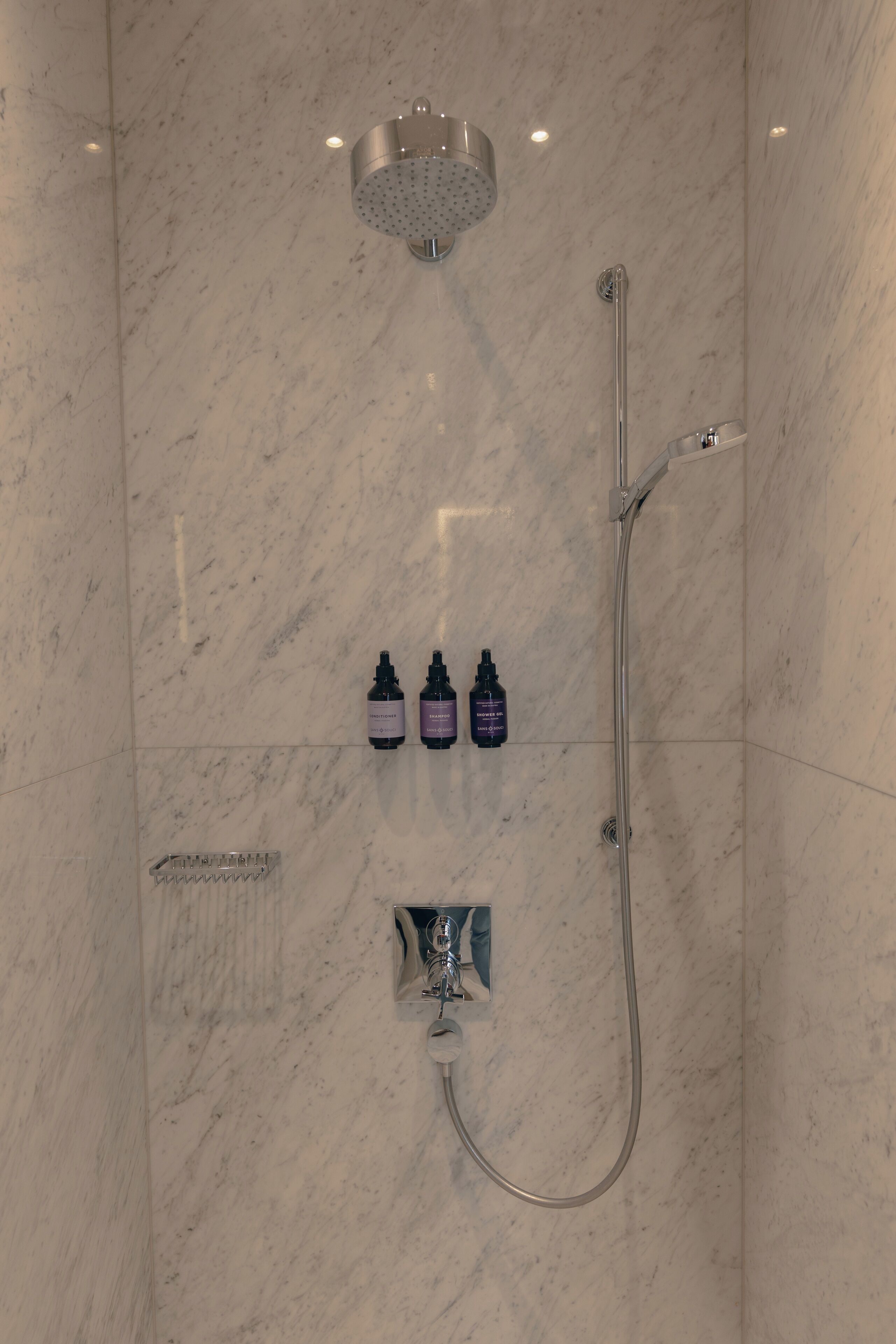 A shower with a handheld showerhead and a wall-mounted faucet. There are three bottles of shampoo and conditioner on the wall.