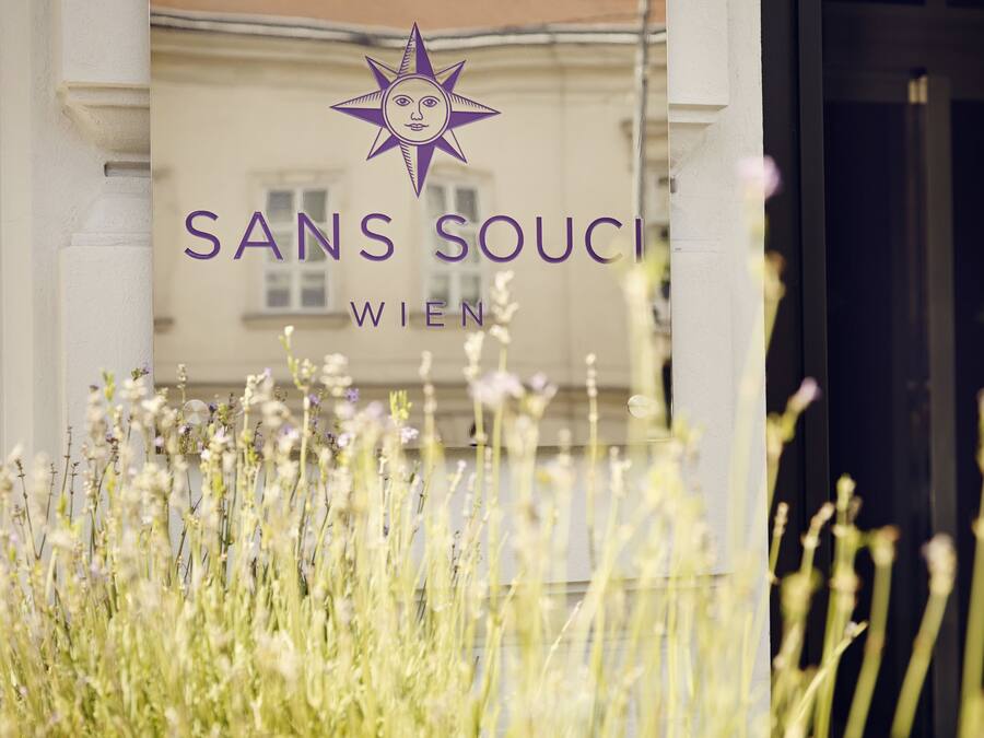 Een bord met 'SANS SOUCI WIEN' en een sterlogo, omringd door begroeiing.