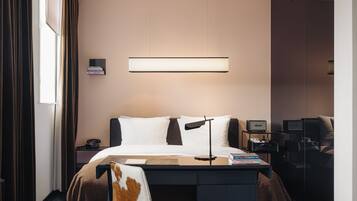 Una habitación de hotel moderna con cama, escritorio, silla y mesita de noche con lámpara y libros.