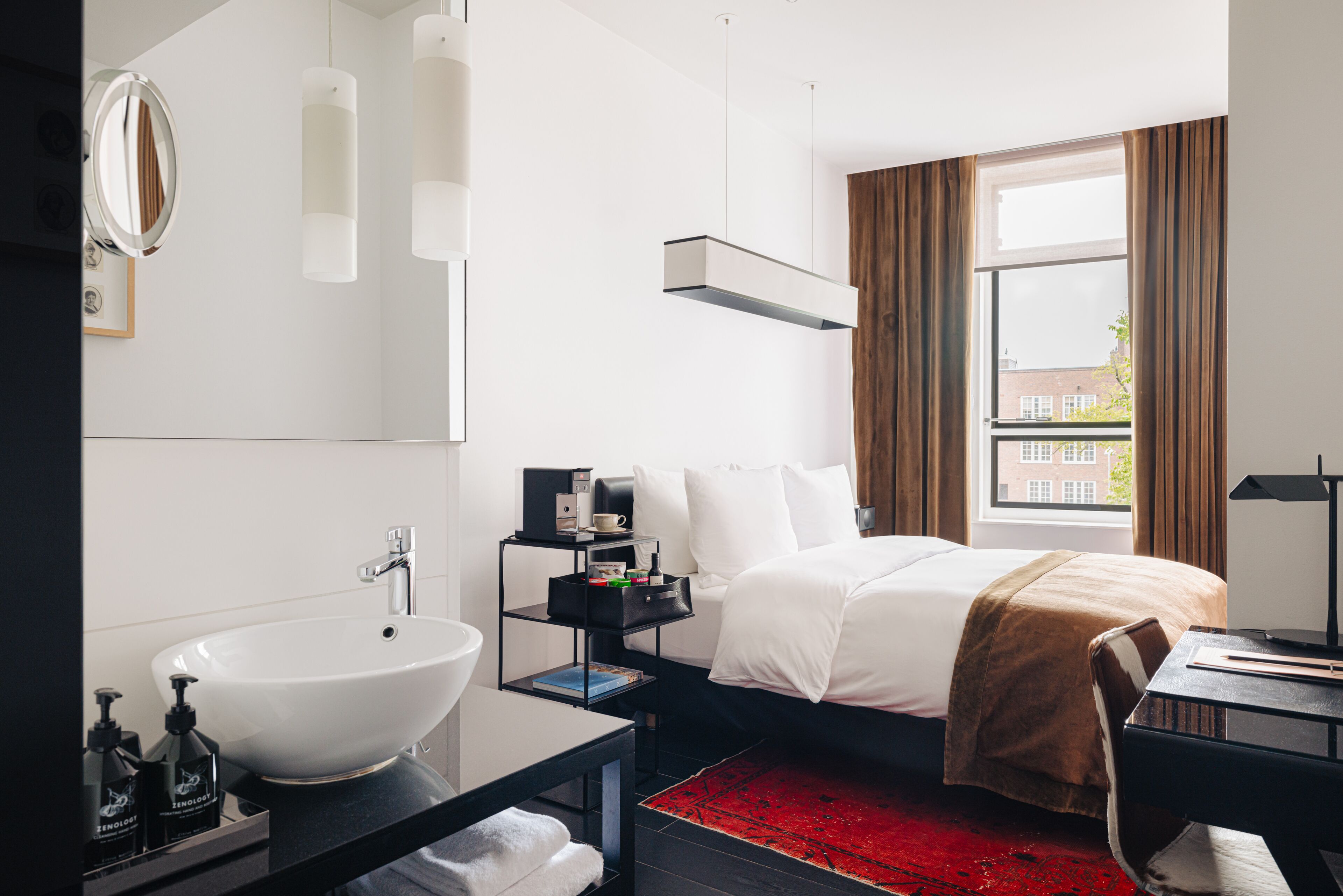 Una habitación de hotel moderna con una cama grande, una mesita de noche con lámpara, un escritorio pequeño, una alfombra roja, un lavamanos blanco y una ventana con cortinas.