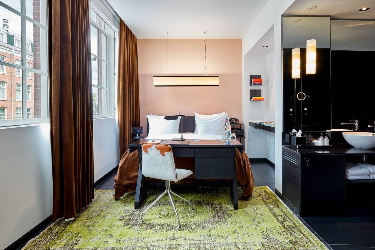 A modern hotel room with a bed, desk, chair, and a bathroom with a sink and mirror.