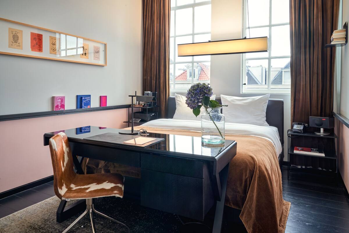 A hotel room with a bed, a desk, a chair, a window with curtains, a bedside table, and a wall-mounted shelf with books.
