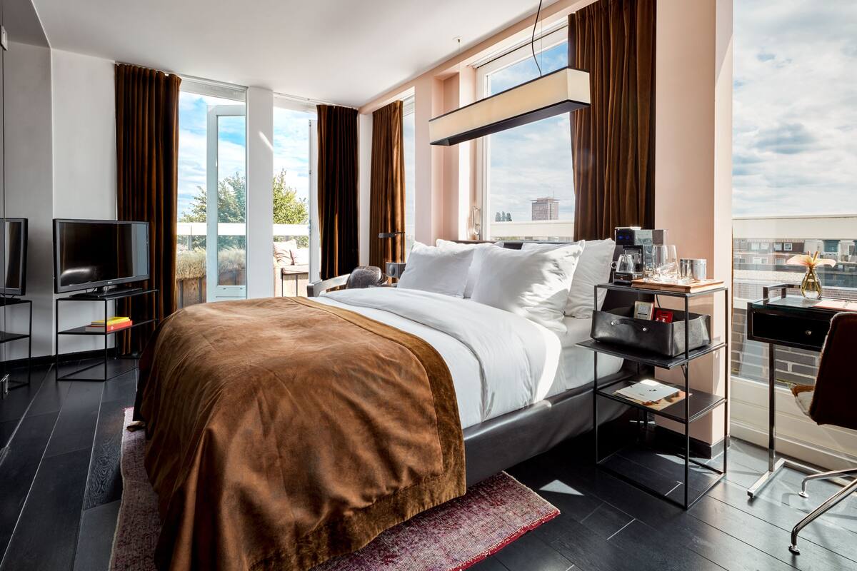 A modern hotel room with a large bed, a desk with a computer, a television, and a view of the city through large windows.