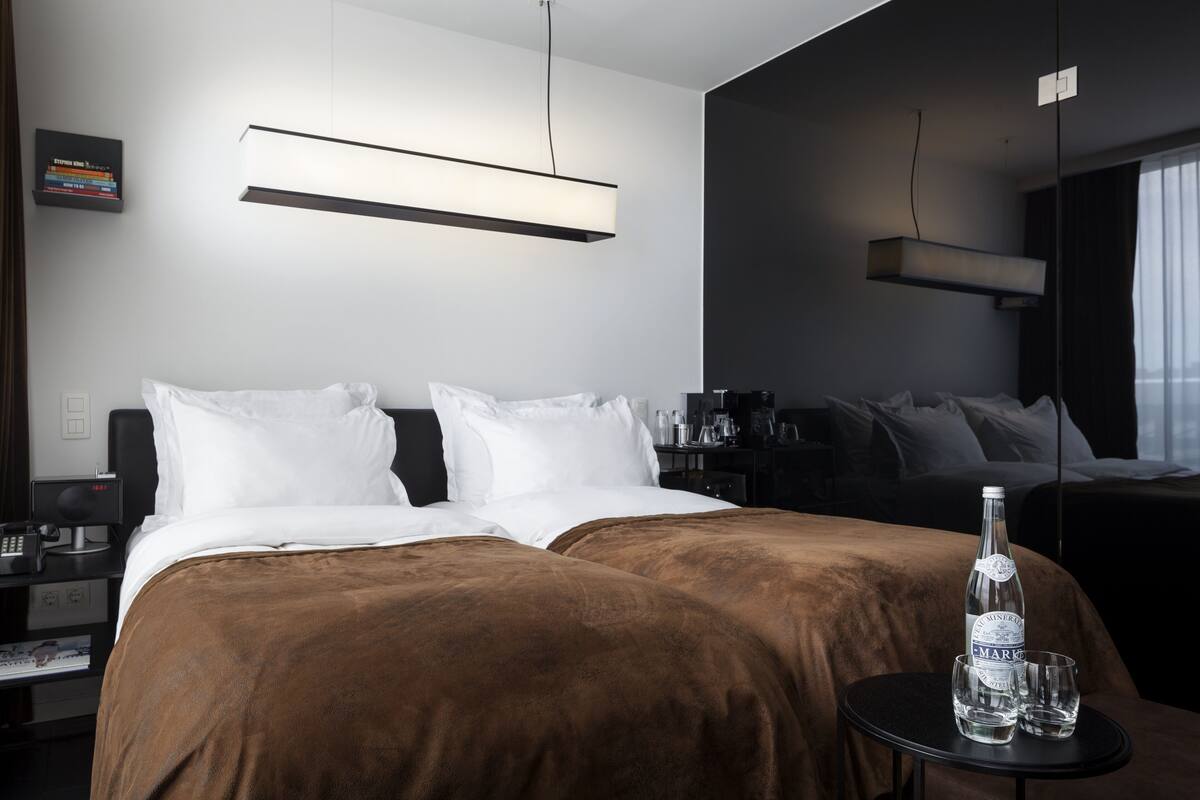 A hotel room with two beds, a bedside table with a bottle and glasses, a desk with a computer, and a wall-mounted shelf with books.
