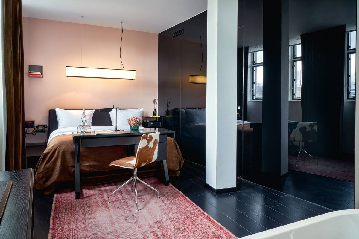 A modern hotel room with a bed, desk, chair, and a view of the city through large windows.