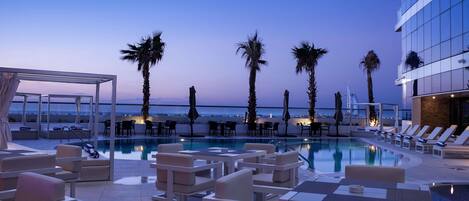 A rooftop pool area with lounge chairs and tables, palm trees, and a view of the city skyline.