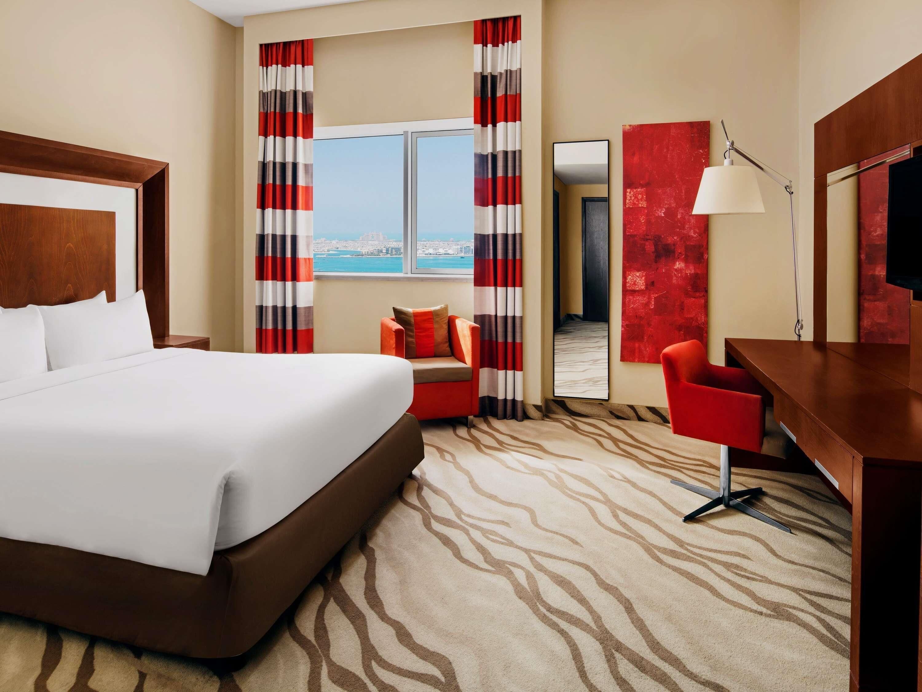 A hotel room with a large bed, a desk, a chair, a mirror, and a view of the city.