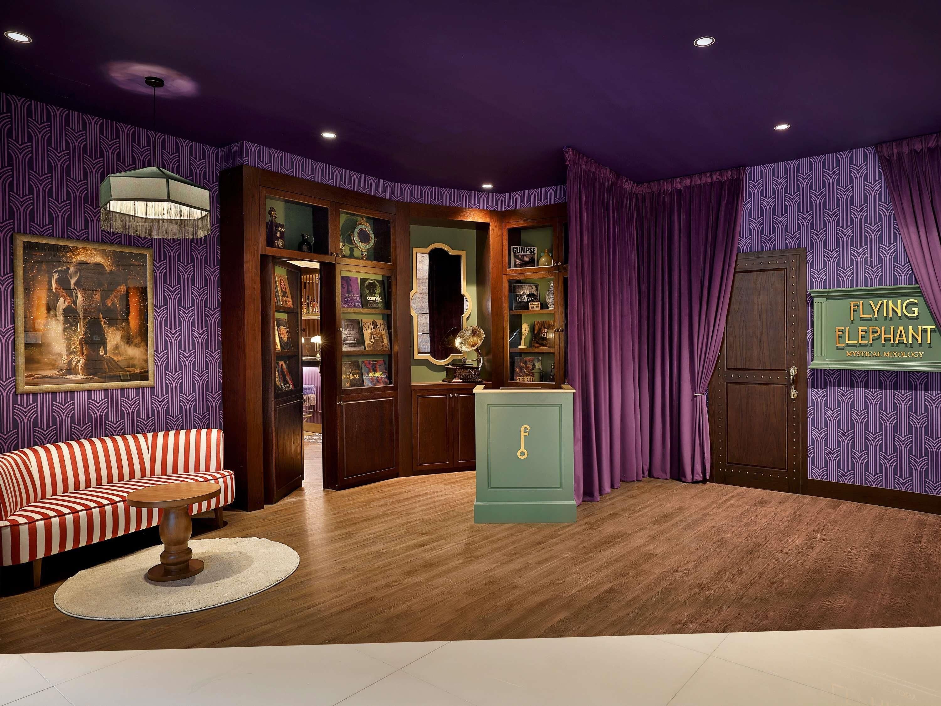 A room with a patterned wallpaper, a sofa, a round table, a cabinet, and a sign reading 'FLYING ELEPHANT MYSTICAL MIXOLOGY'.