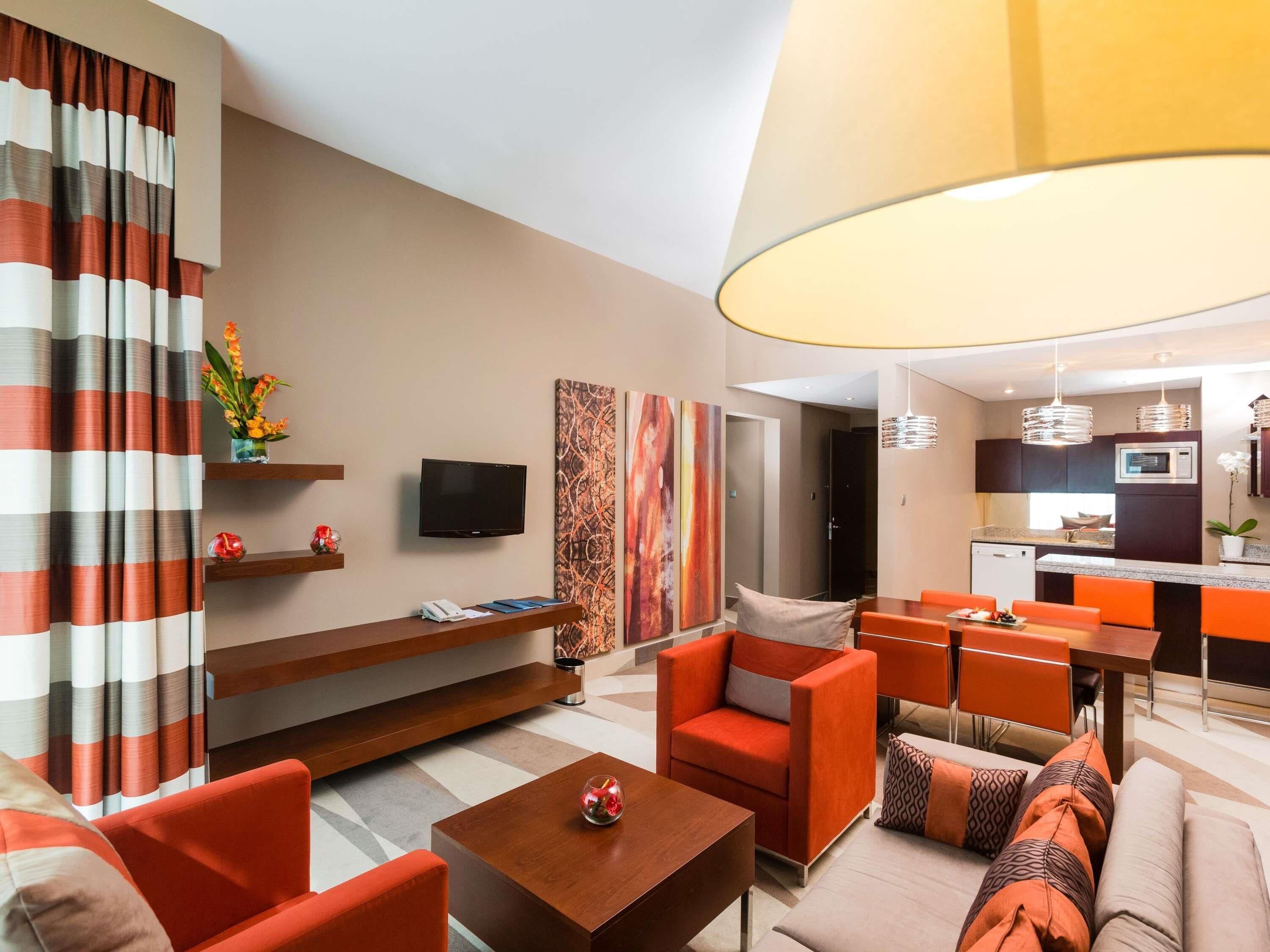 A modern living room with a sofa, chairs, a coffee table, and a TV. There is a kitchen area with a dining table and chairs.