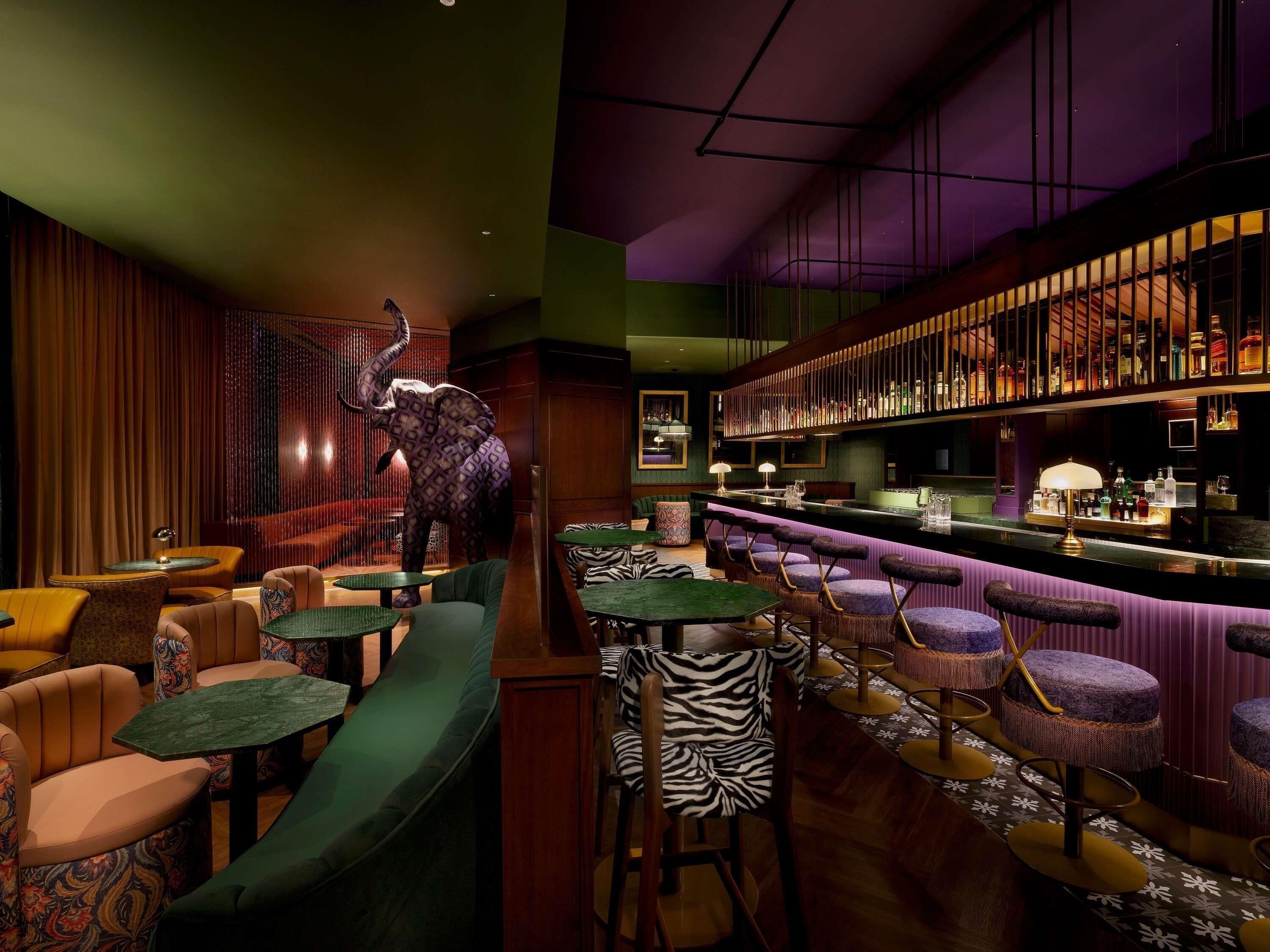 A bar area with a large elephant statue, multiple seating options including bar stools and cushioned chairs, and a well-stocked bar counter.