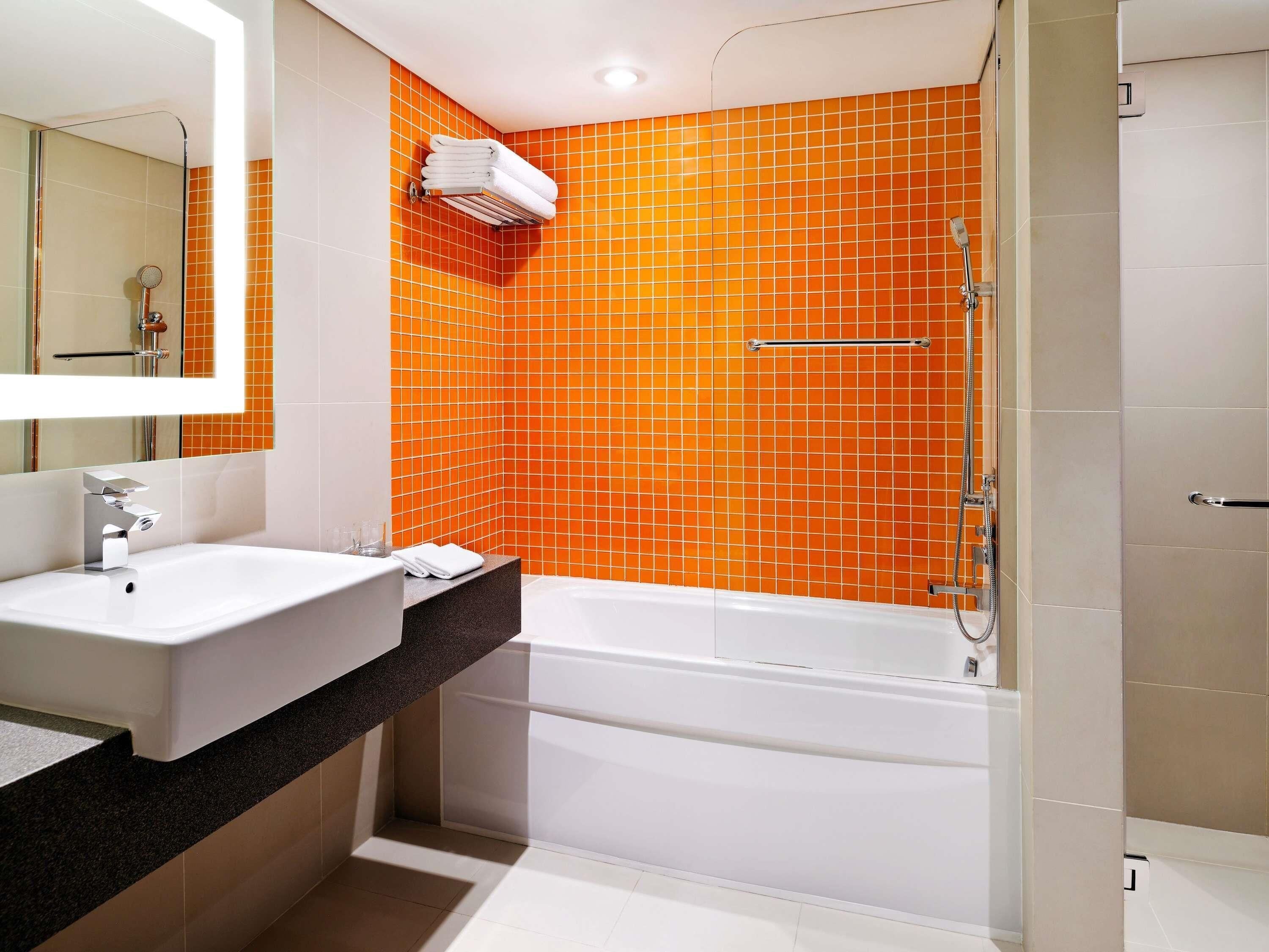 A modern bathroom with a white sink, a large mirror, an orange-tiled shower wall, and a white bathtub.