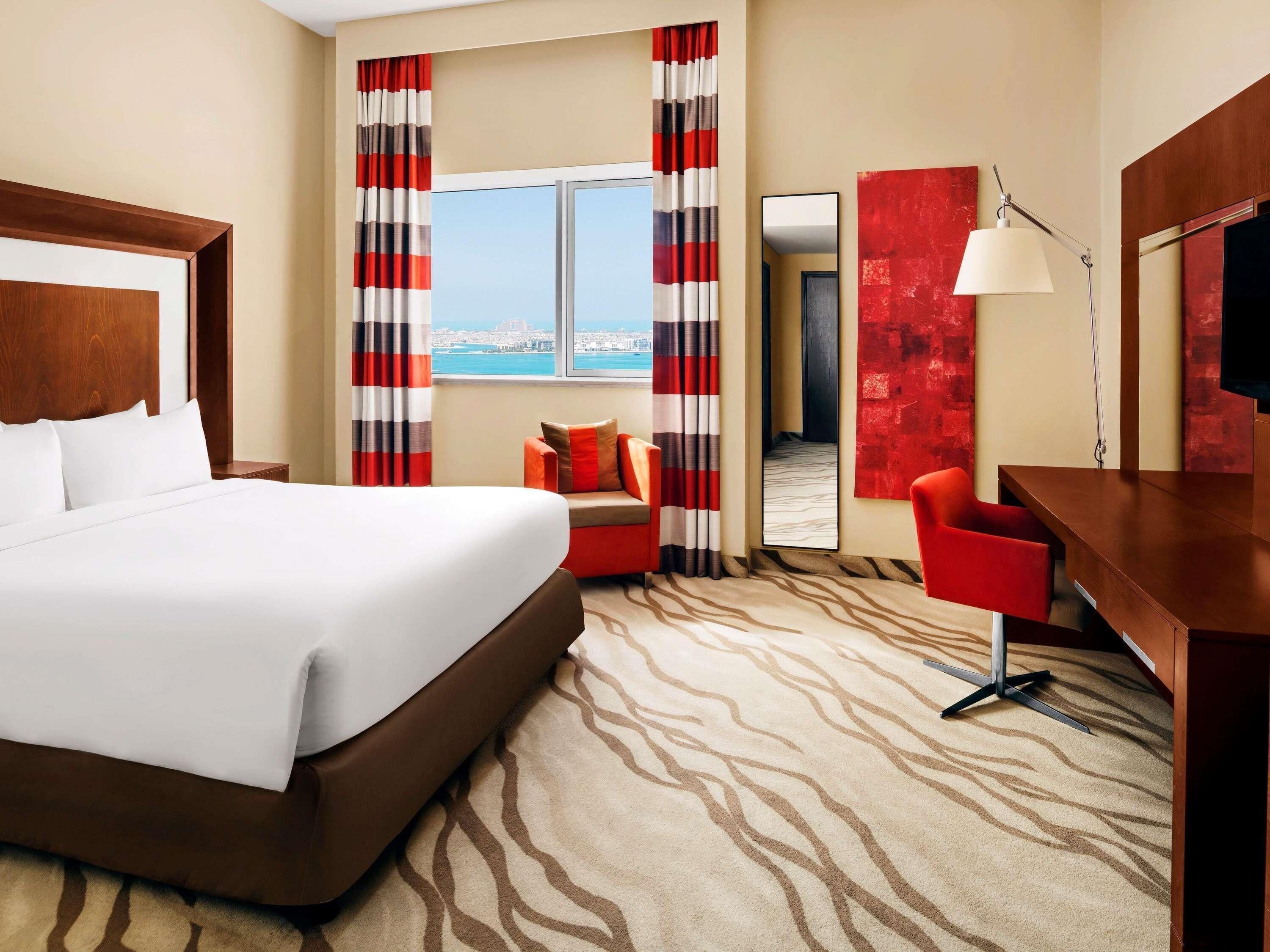 A hotel room with a large bed, a desk, a chair, a lamp, and a view of the city.