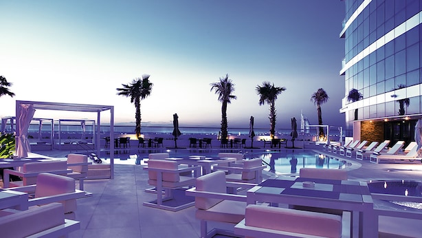 Área de piscina en la azotea con sillones de descanso, palmeras y vistas de la ciudad.