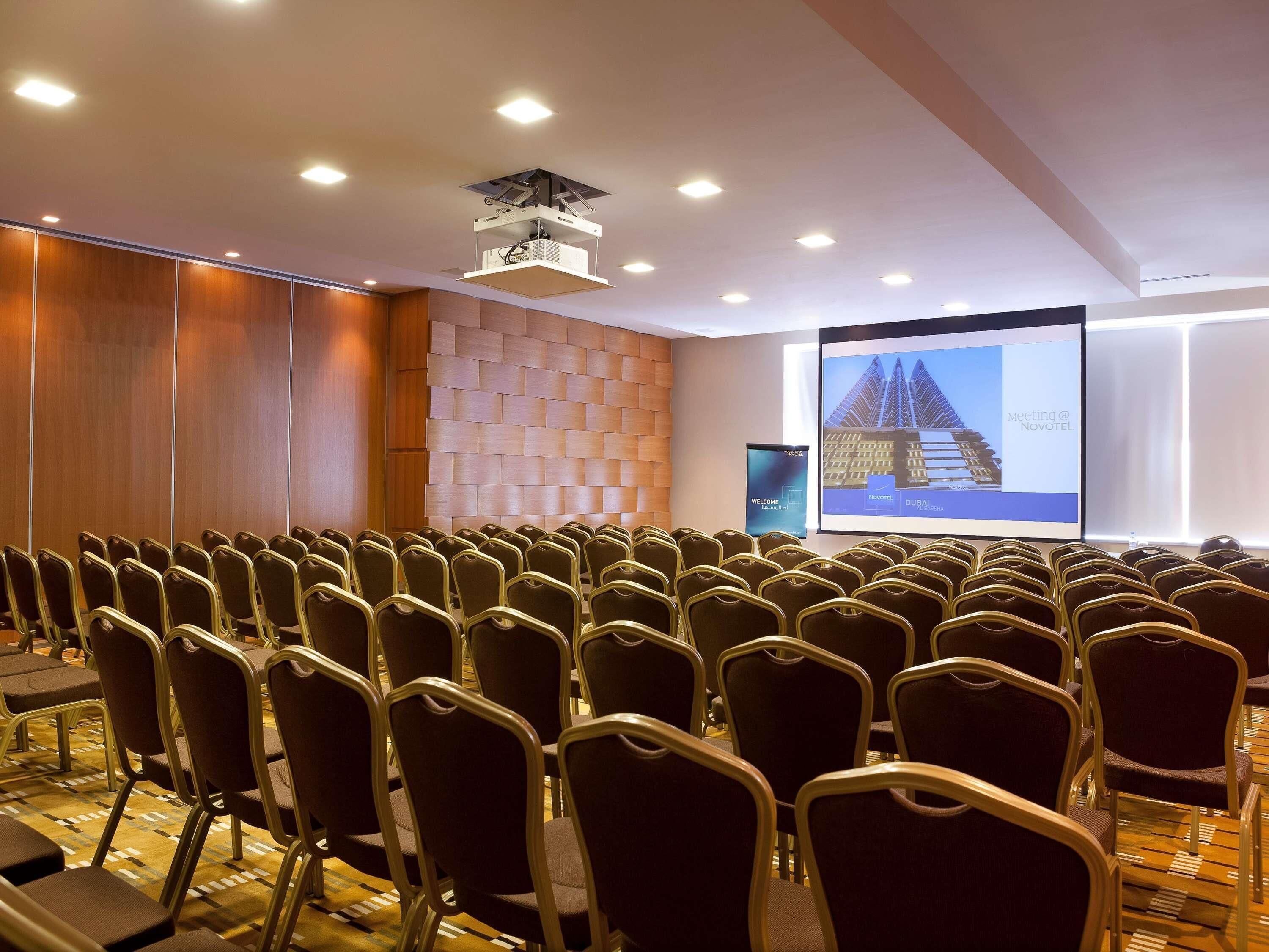 A conference room with rows of chairs, a projector screen, and a wooden panel wall.
