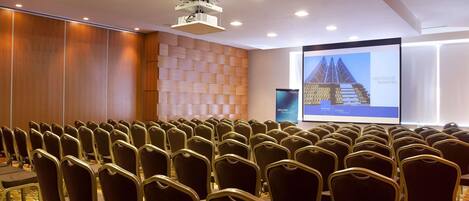 A conference room with rows of chairs, a projector screen, and a wooden panel wall.