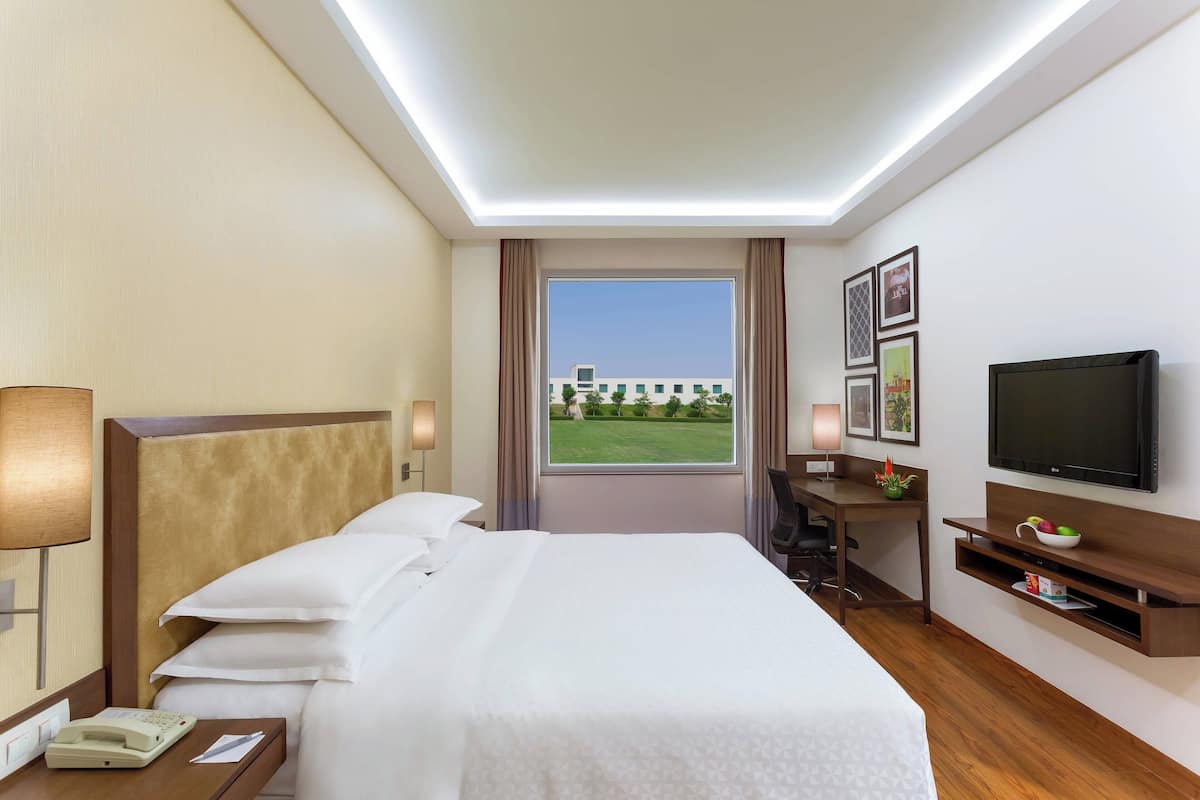 Ein Hotelzimmer mit Bett, Nachttisch, Lampe, Schreibtisch, Fernseher und einem Fenster mit Blick auf ein Gebäude und Grünflächen.