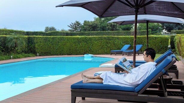 A man relaxing on a sun lounger by a pool with an umbrella, surrounded by neatly trimmed hedges and other loungers. - Four Points by Sheraton New Delhi, Airport Highway (New Delhi)