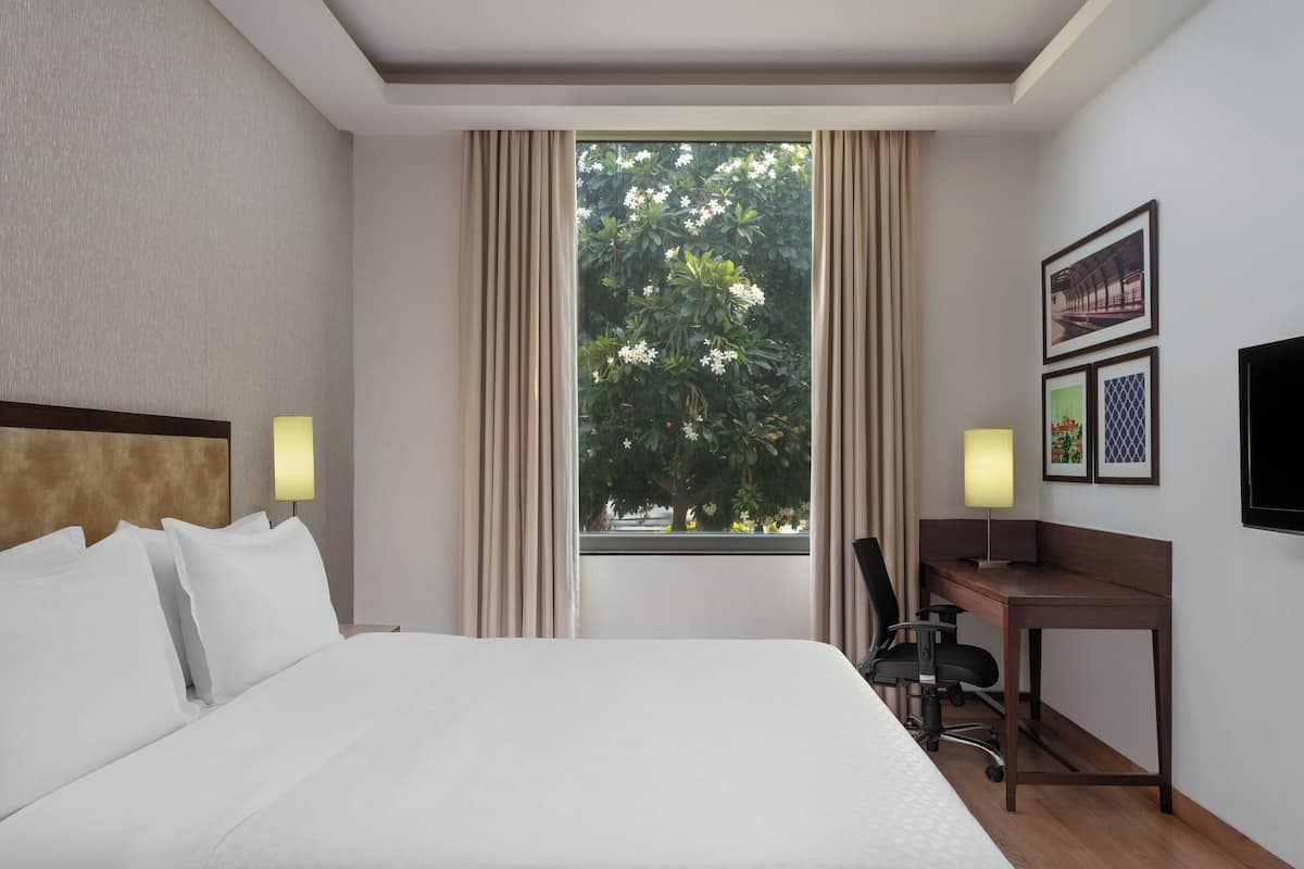 Ein Hotelzimmer mit Bett, Schreibtisch, Stuhl, einem fensterbelaubten Fenster und Wandbildern.