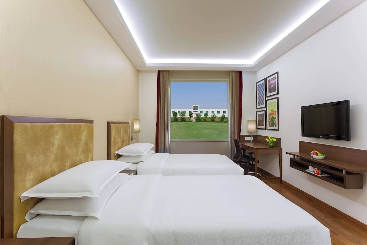 Ein Hotelzimmer mit zwei Betten, einem Fernseher, einem Schreibtisch und einem Fenster mit Blick auf ein Gebäude.