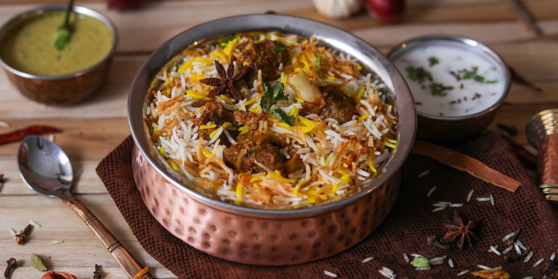 A copper bowl of biryani with meat and rice, surrounded by spices and a spoon.