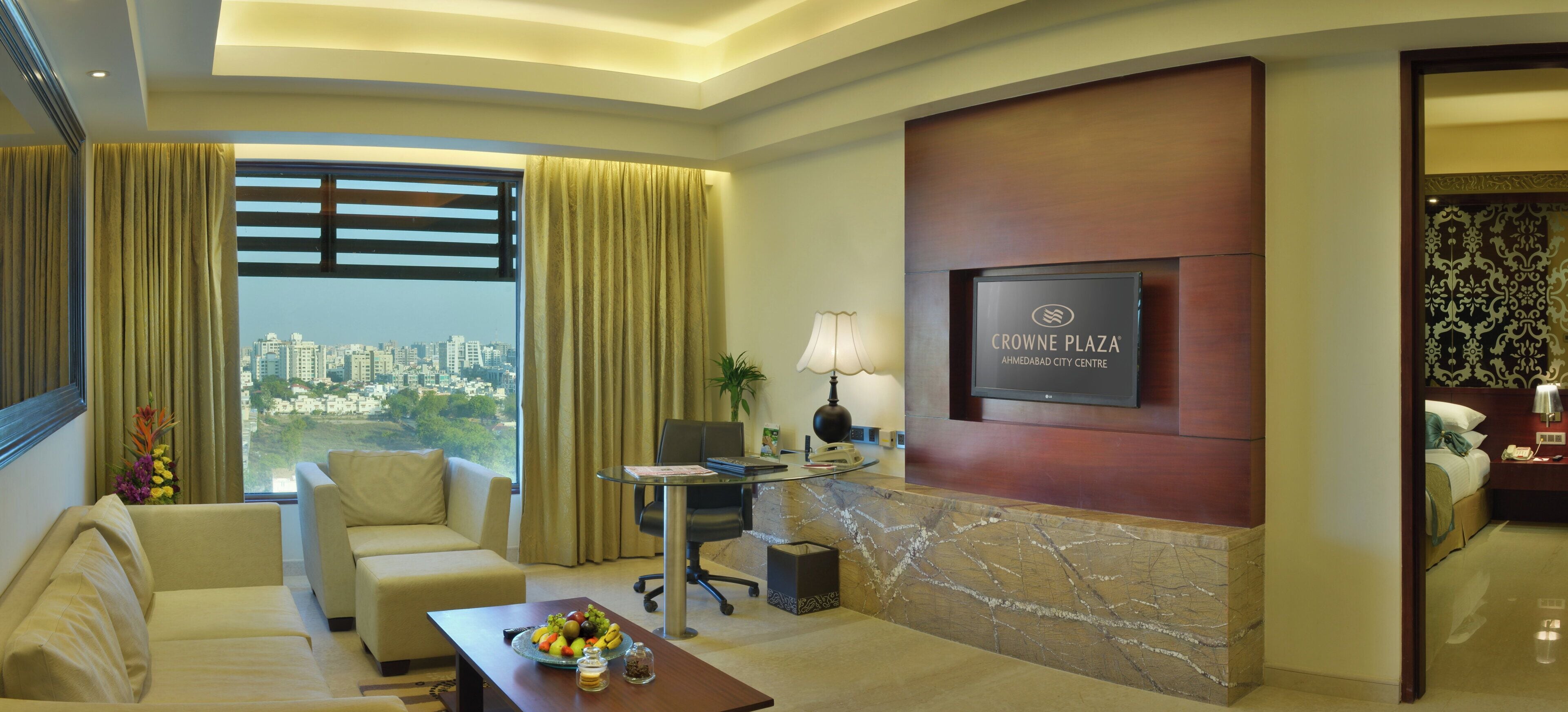 A hotel room with a city view, a flat-screen TV, a desk with a lamp, and a bed.