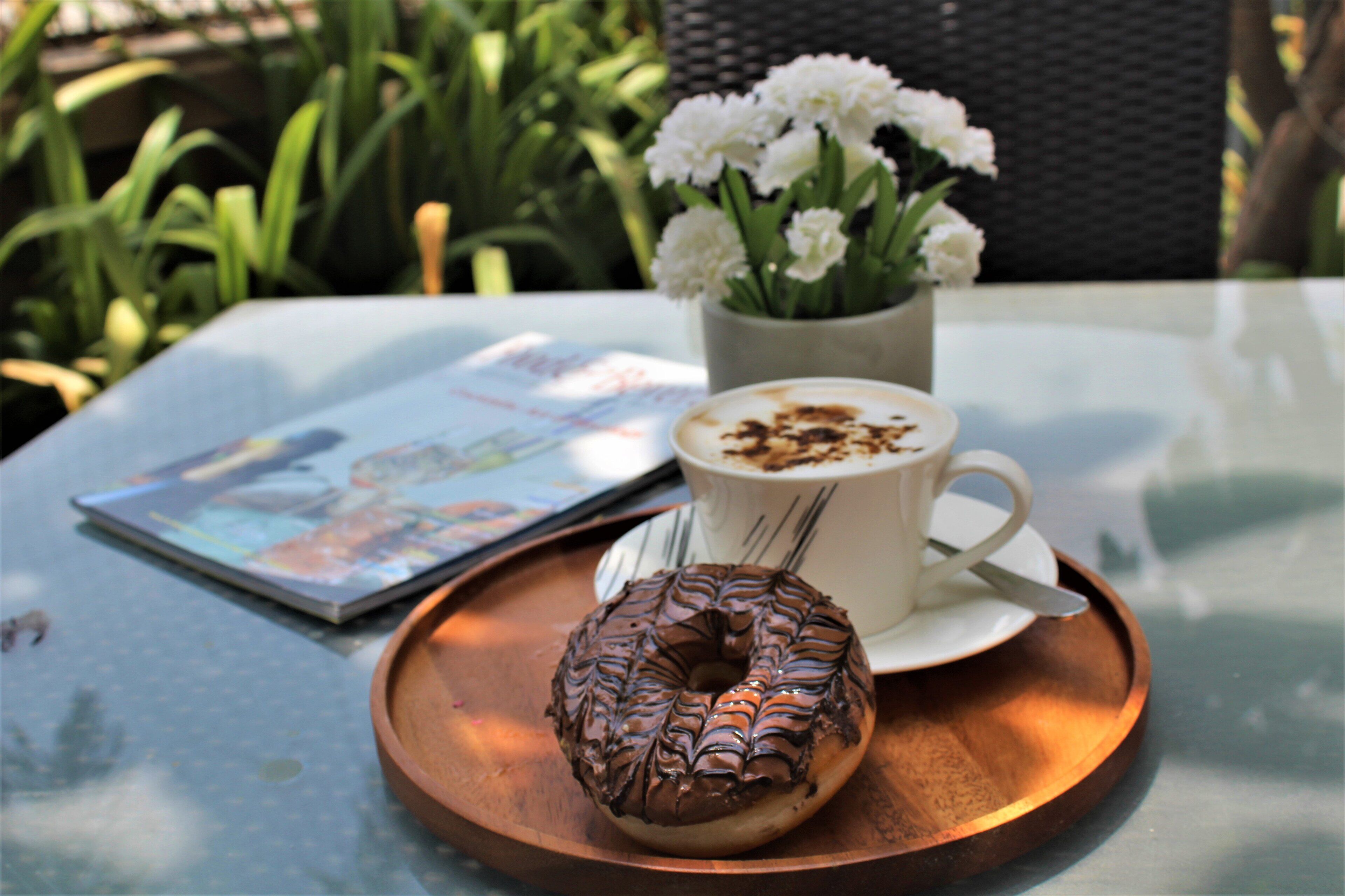A table with a tray holding a chocolate donut and a cup of coffee, accompanied by a magazine and a vase of white flowers.