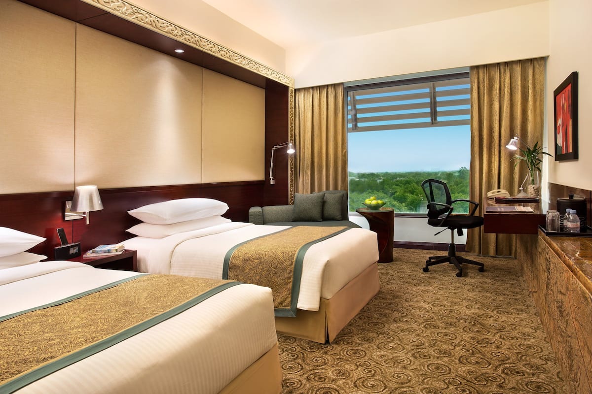 Ein Hotelzimmer mit zwei Betten, einem Schreibtisch, einem Stuhl, einer Schreibtischlampe und Blick auf Grün durch das Fenster.