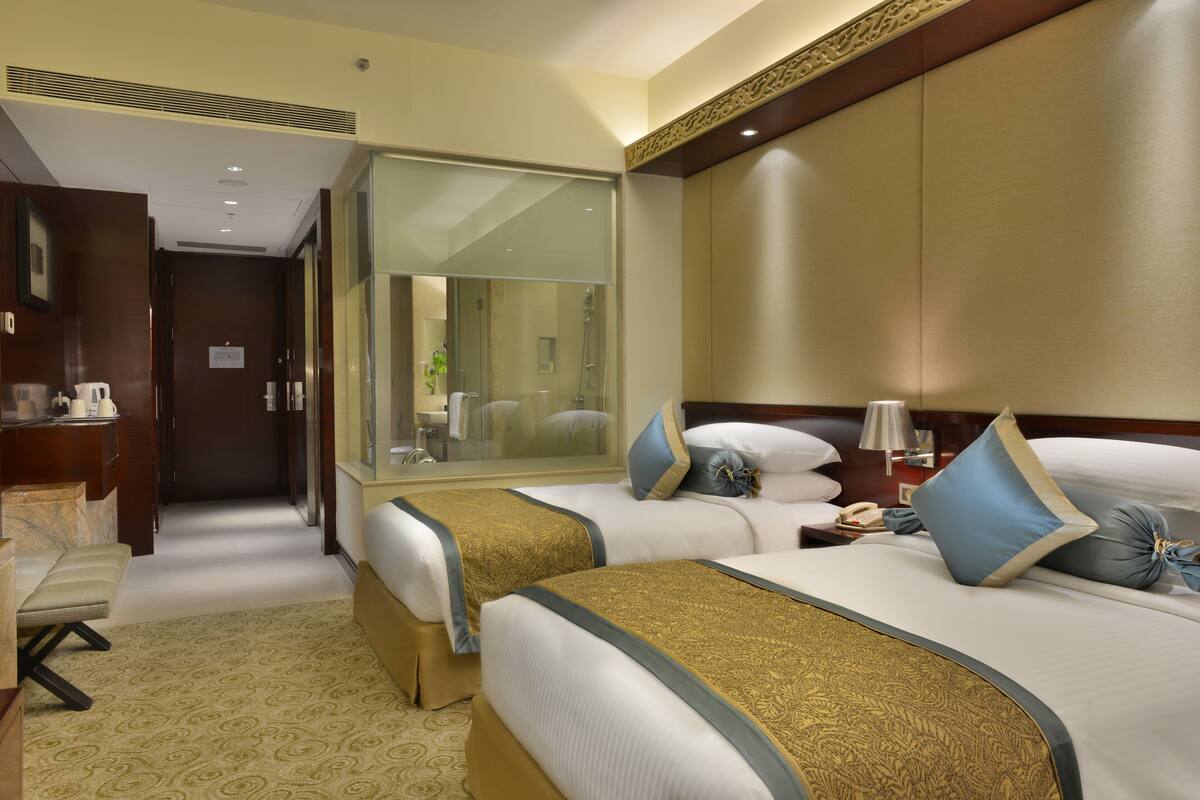 Ein Hotelzimmer mit zwei Betten, einem Schreibtisch, einem Spiegel und einem durch eine Glastür einsehbarer Badezimmer.