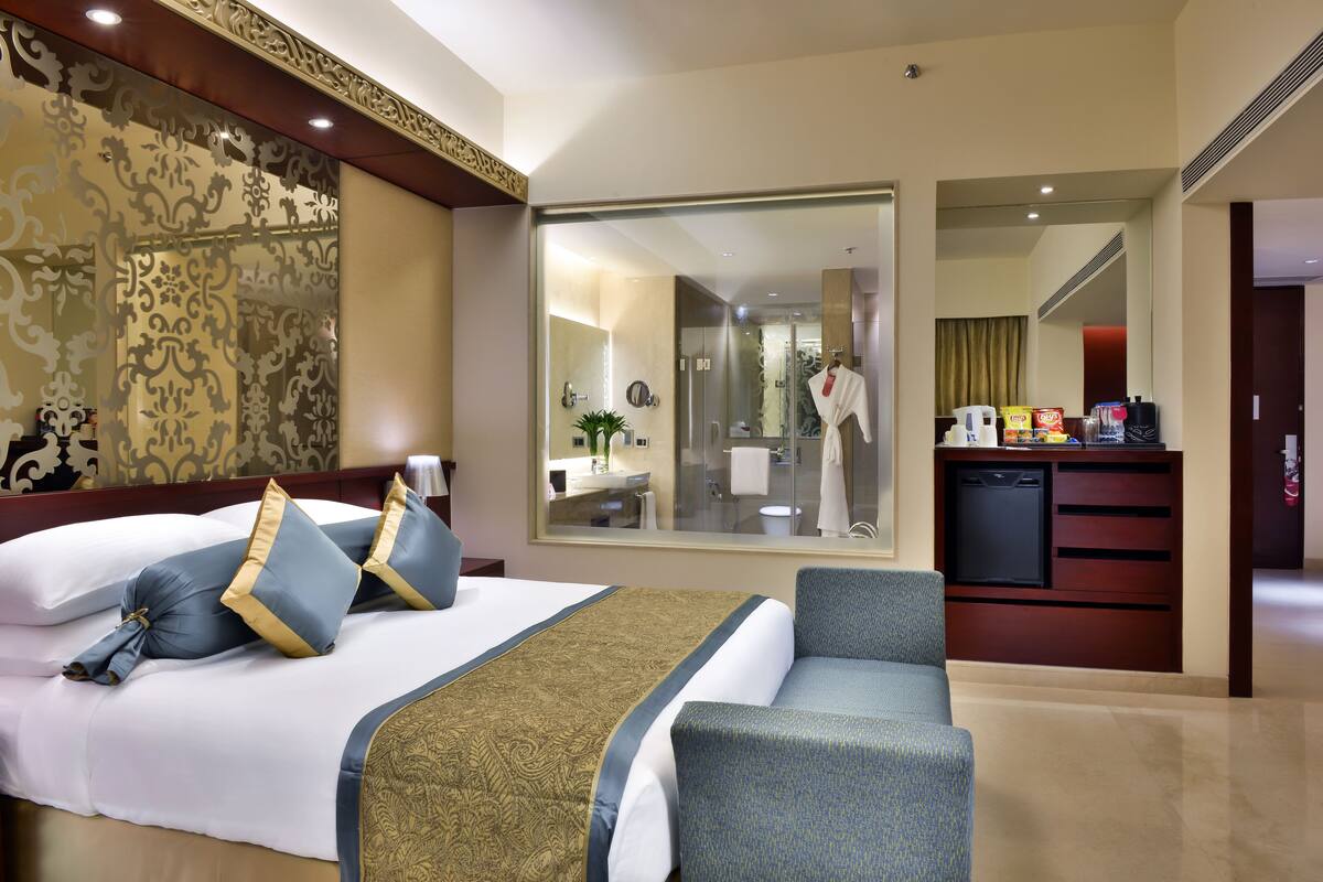 Ein Hotelzimmer mit einem großen Bett, einer Sitzecke, einem durch eine Glastür einsehbaren Badezimmer und einer Küchenzeile.