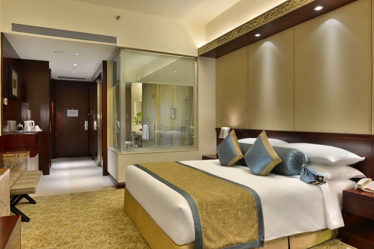 Ein Hotelzimmer mit einem großen Bett, einem Nachttisch, einem Spiegel, einem Fenster und einem durch eine Glastür einsehbarer Badezimmer.