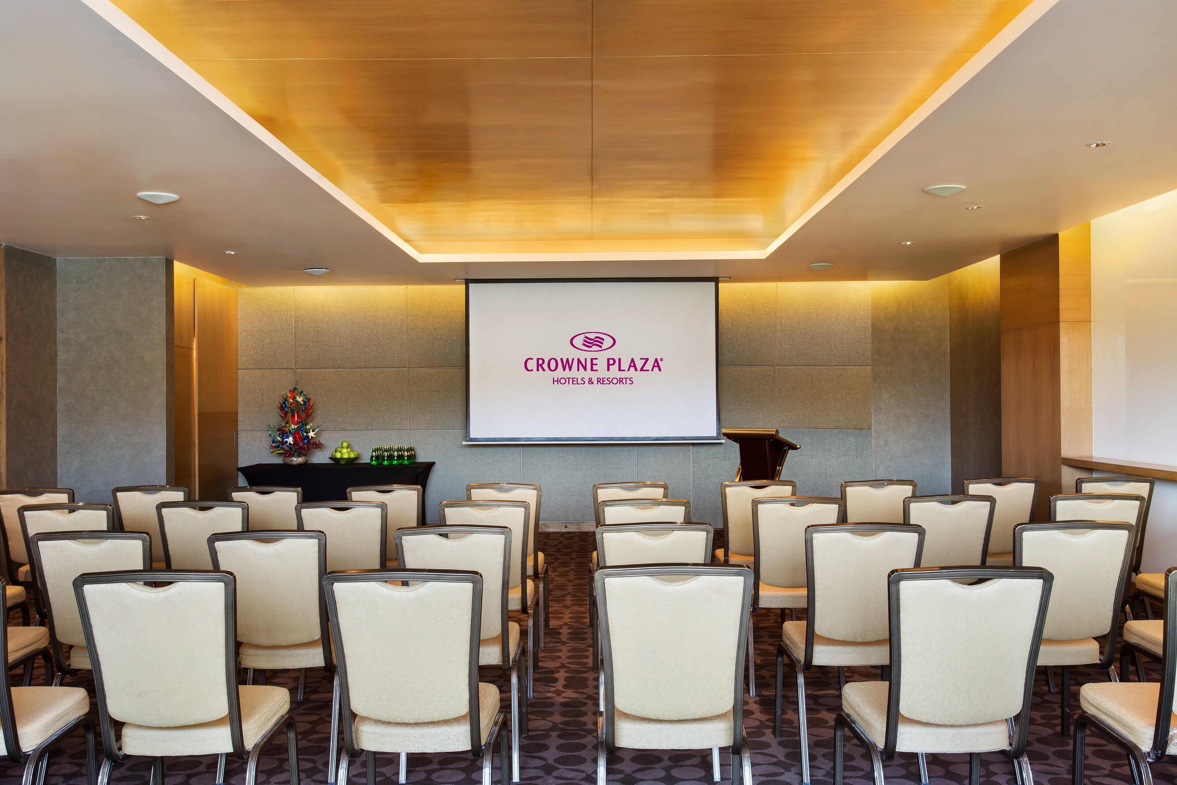 Un salón de conferencias con filas de sillas apuntando hacia una pantalla de proyección que muestra el logotipo de Crowne Plaza Hotels & Resorts.
