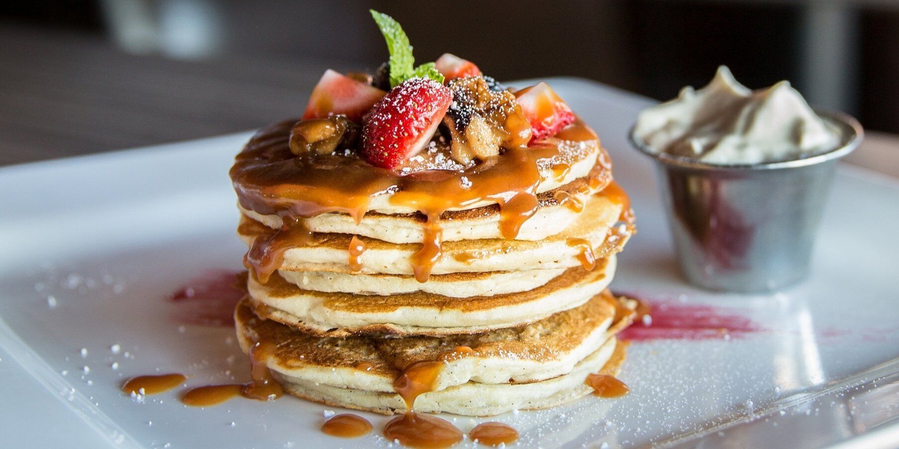 A stack of pancakes with caramel sauce and fruit toppings, served on a white plate with a small cup of whipped cream.