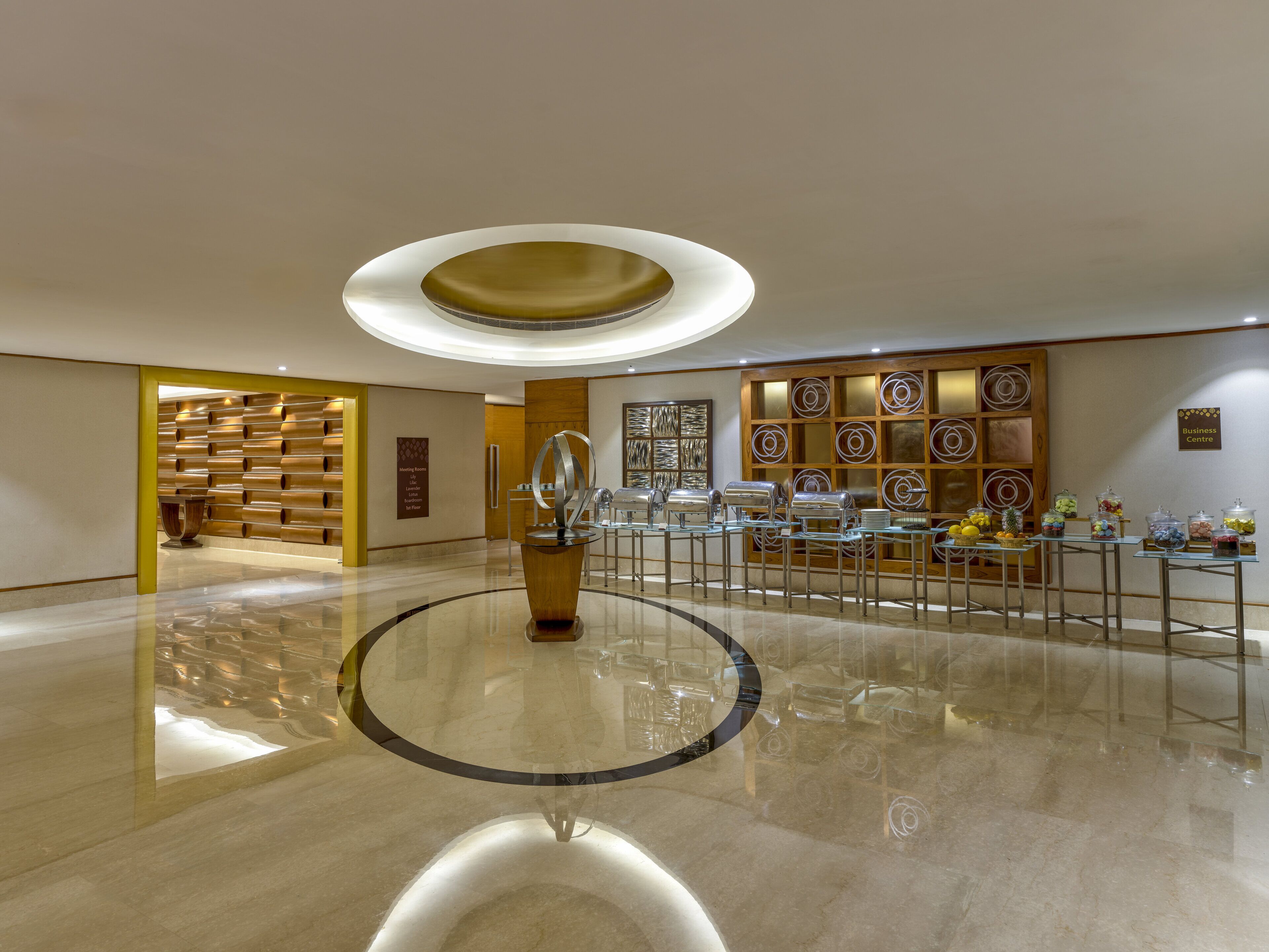 A modern hotel lobby with a central glass table, a meeting room sign, and a business center sign.
