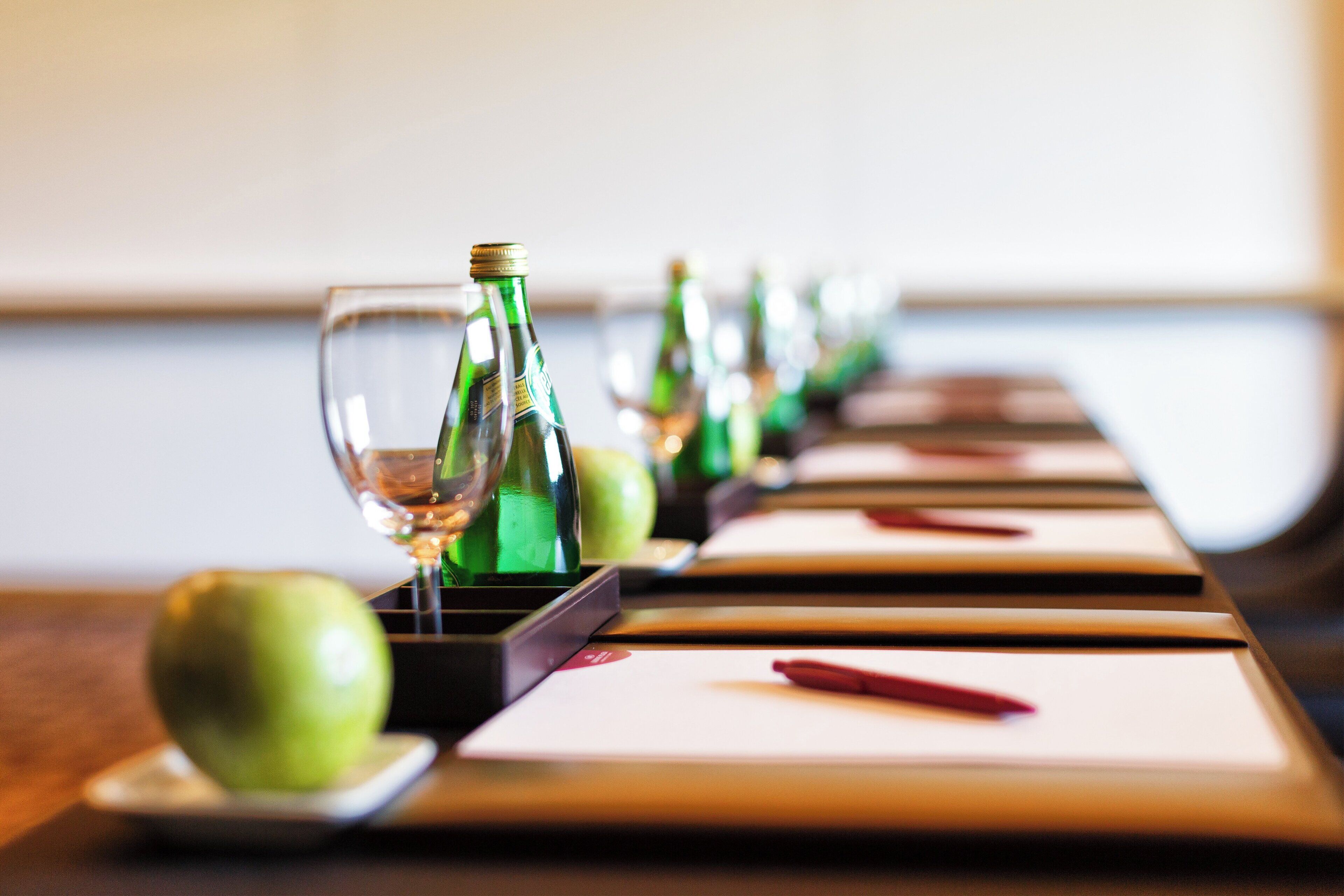 A long table with wine glasses and bottles, apples, and red pens.
