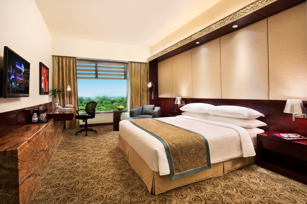 Ein Hotelzimmer mit einem großen Bett, einem Schreibtisch mit Computer, einem Sessel und Blick ins Freie.