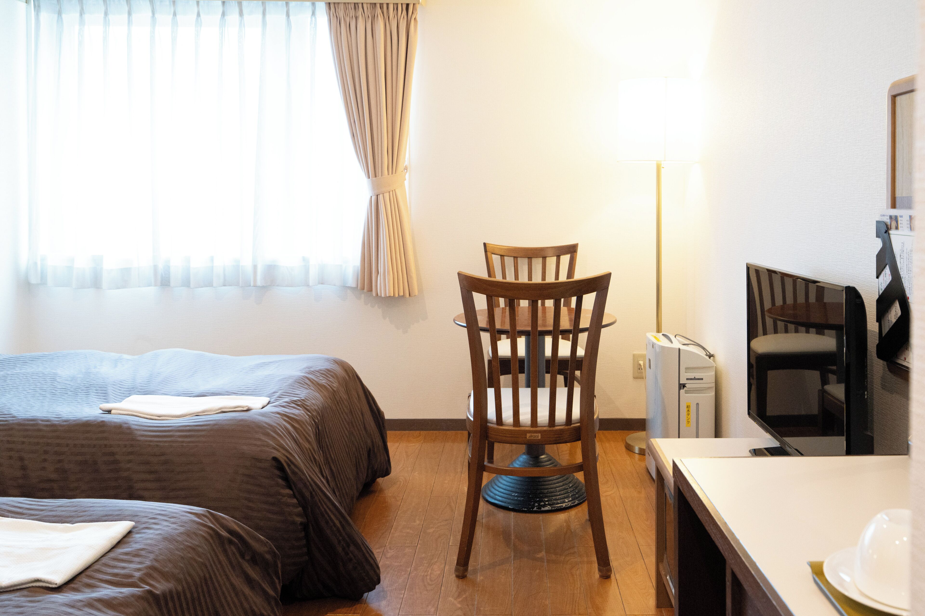 Une chambre d’hôtel avec deux lits, une chaise en bois, un bureau, un réfrigérateur et une lampe.