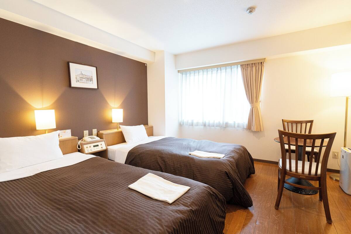 Habitación de hotel con dos camas, una mesita de noche con teléfono, una silla y una ventana con cortinas.