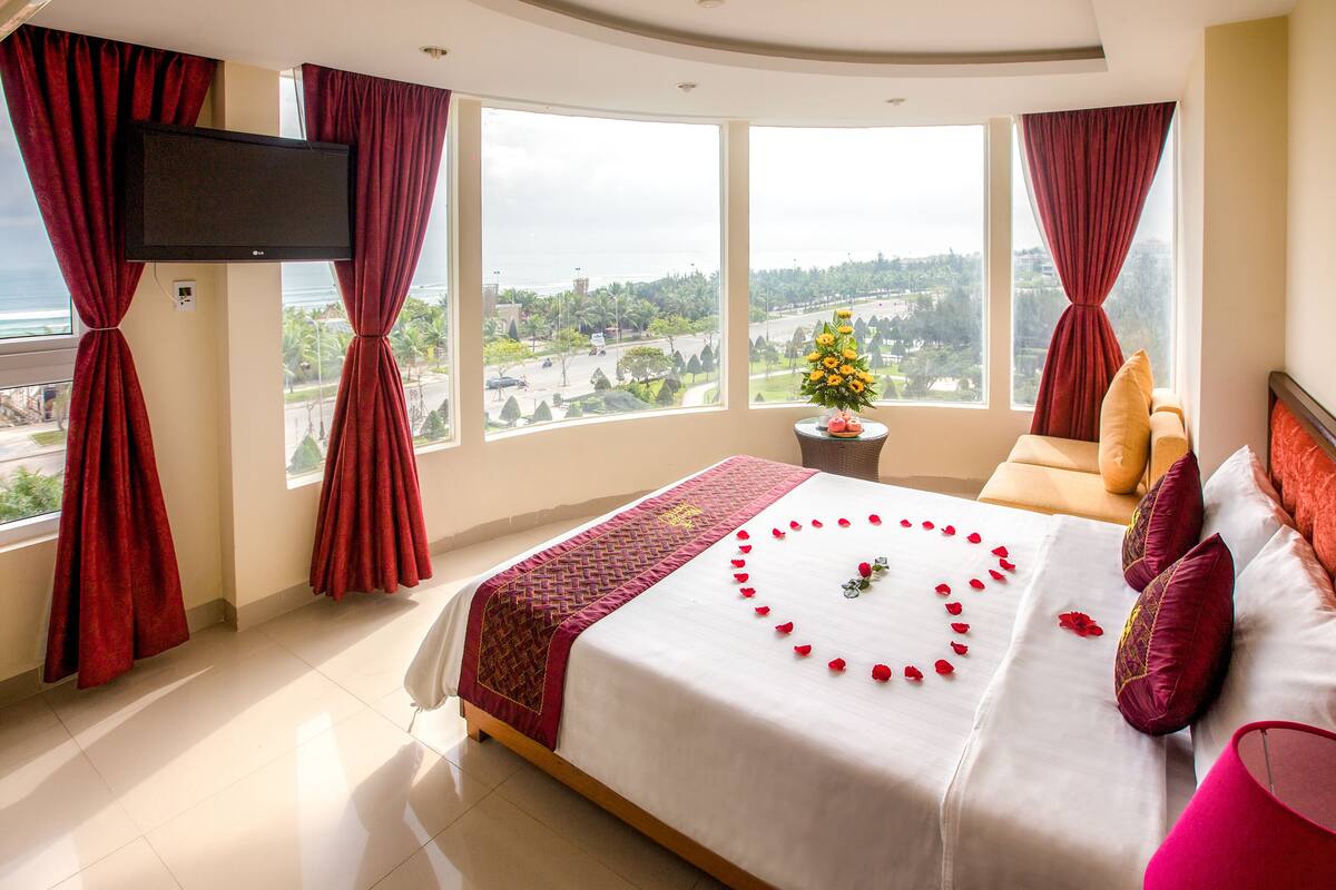 A hotel room with a large bed, red curtains, a TV, a flower arrangement, and a view of a cityscape.