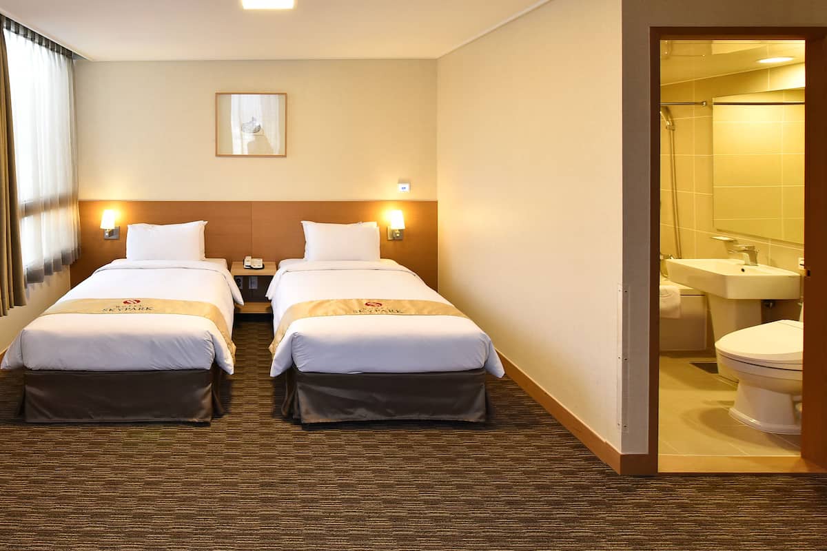 Et hotelværelse med to senge, et natbord med lampe, et billede i ramme på væggen, og et badeværelse, der kan ses gennem en åben dør.