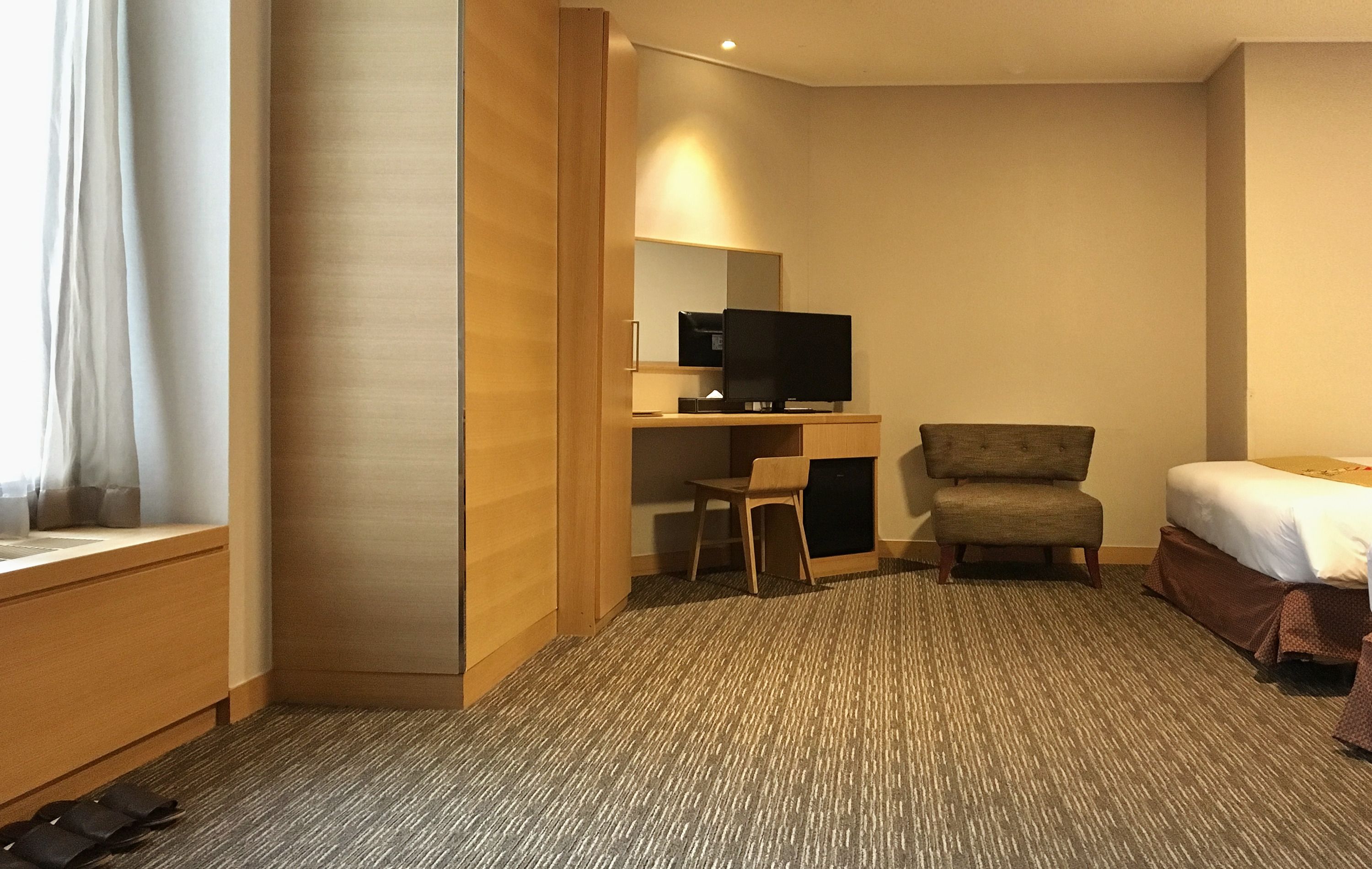 Quarto de hotel com cama, escrivaninha, televisão, cadeira e janela com cortinas.